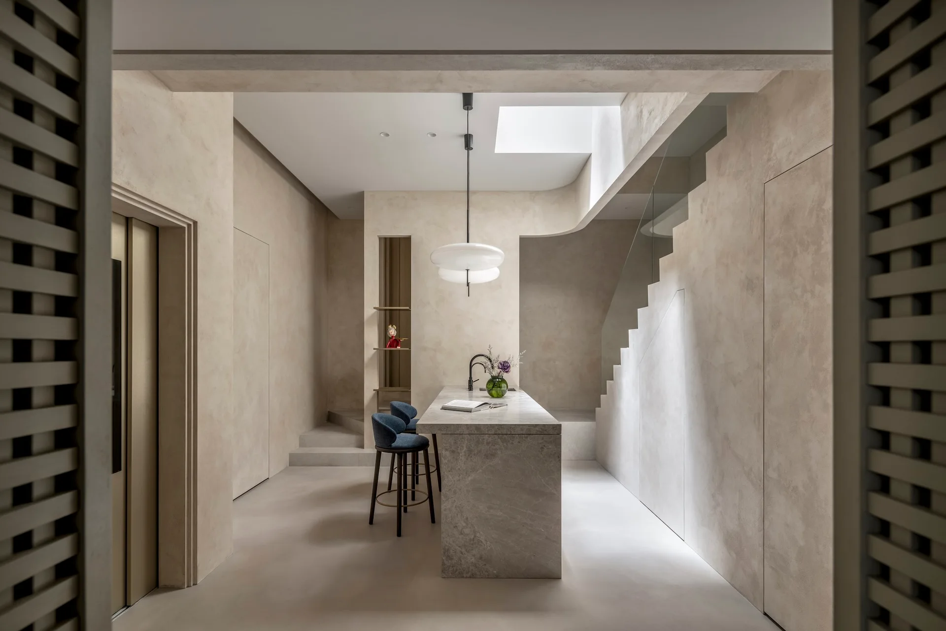 A minimalist kitchen features a stone island with blue bar stools, a sculptural staircase, textured beige plaster walls, and an overhead skylight.