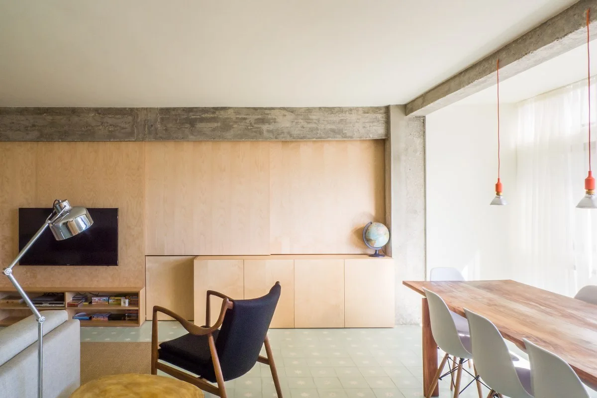 A bright open-plan room featuring light wood wall paneling, an exposed concrete beam, a dining table with white chairs, and a dark armchair.
