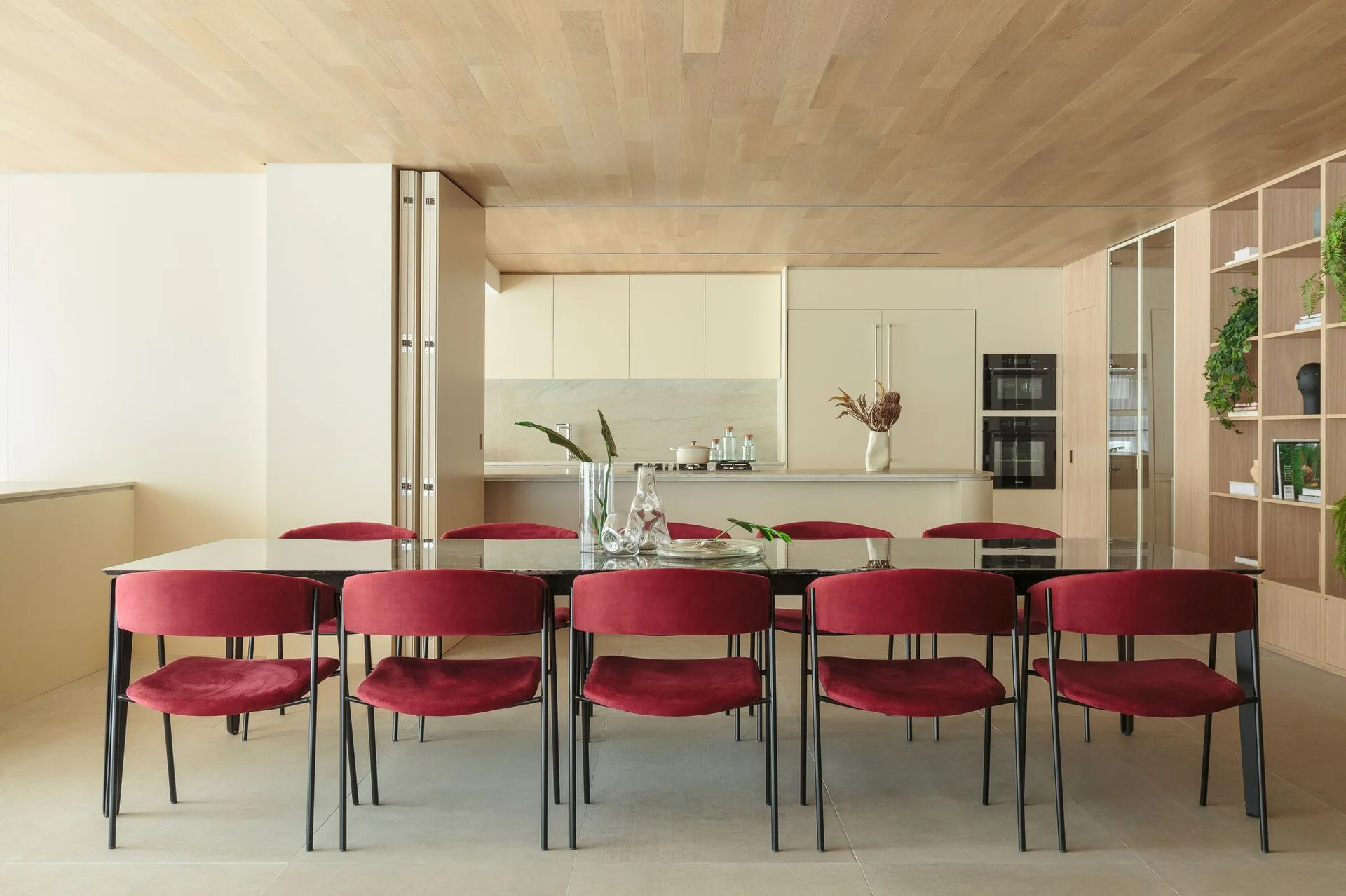 Ten crimson velvet chairs surround a long dark dining table under a light wood paneled ceiling next to a minimalist cream kitchen.