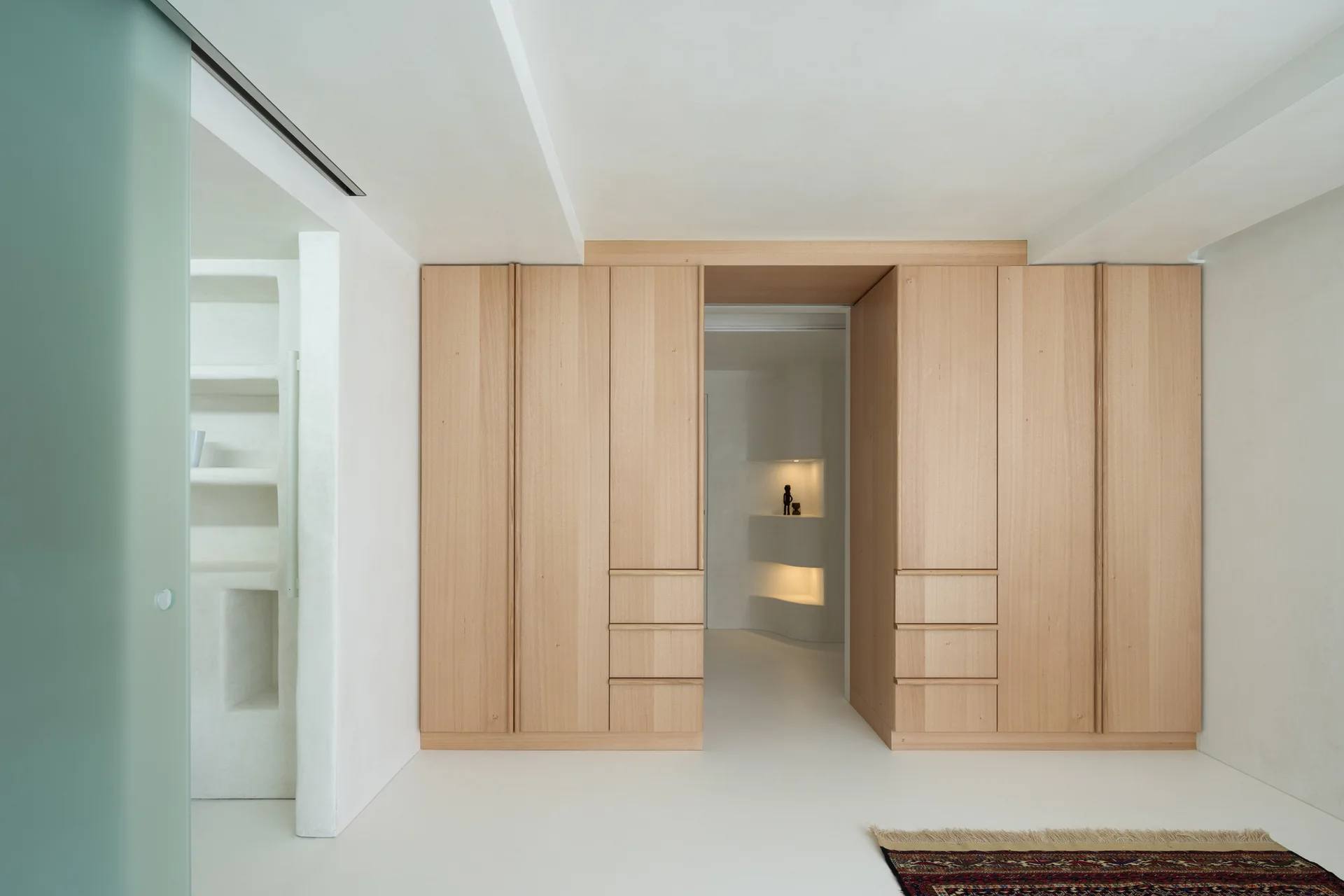 A minimalist dressing room features custom floor-to-ceiling light wood wardrobes framing a passage to an illuminated plaster alcove.