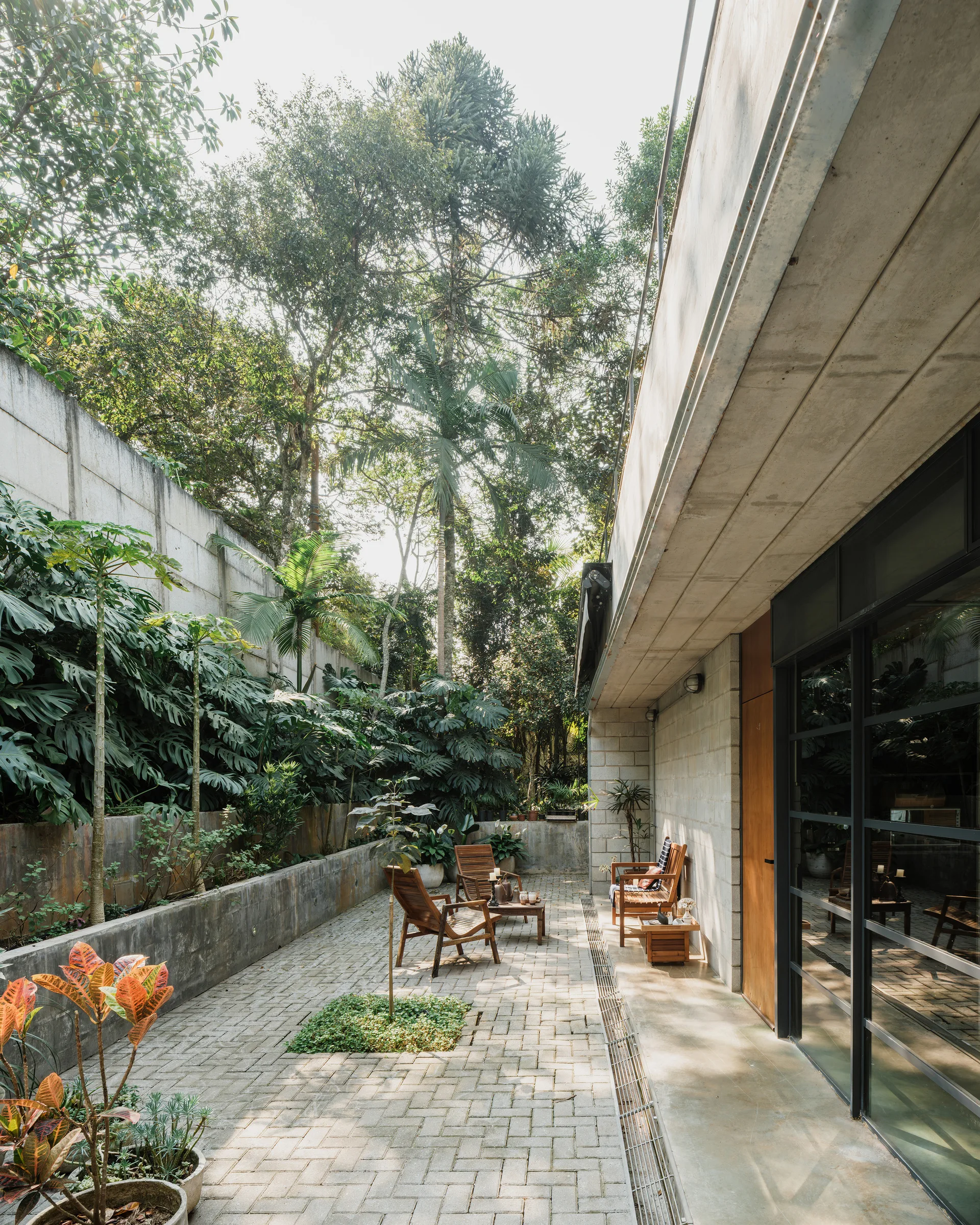 Wooden patio furniture sits on herringbone brick pavers in an outdoor courtyard framed by structural concrete walls and lush tropical greenery.