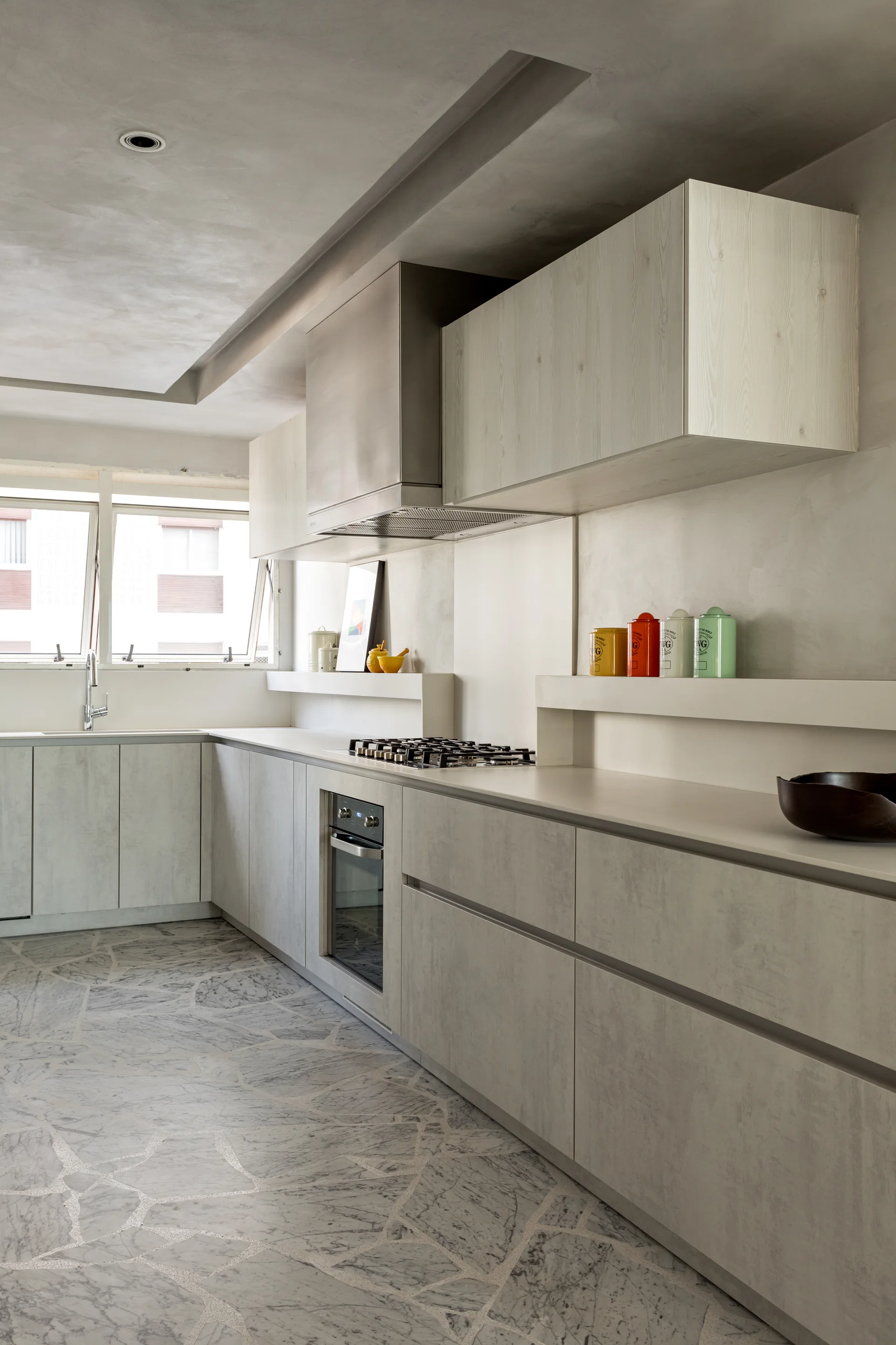 A modern kitchen featuring textured gray plaster walls, light wood upper cabinets, a built-in oven, and a mosaic-style crazed stone tile floor.