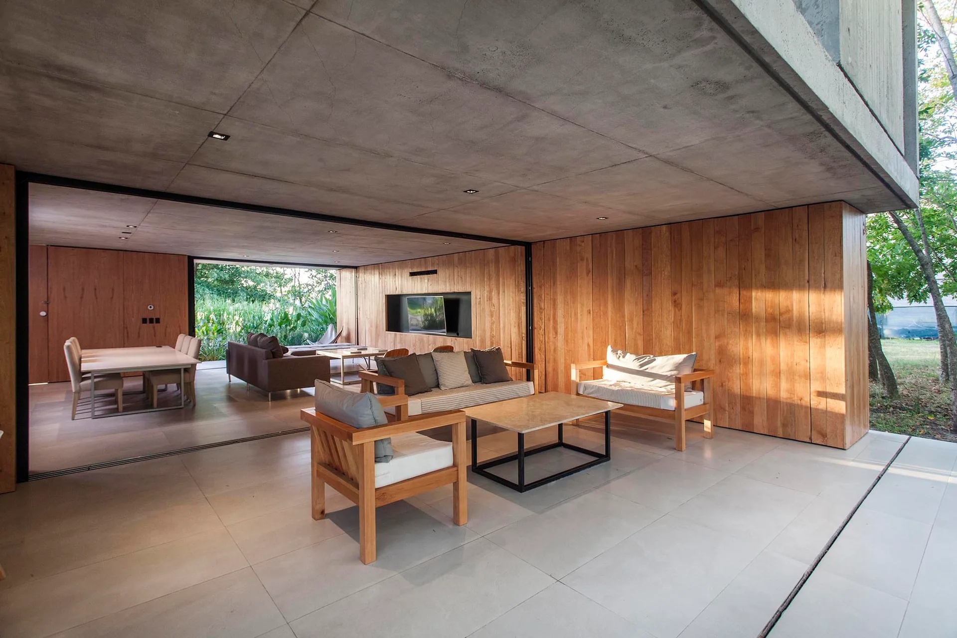 A covered modern patio features a grey concrete ceiling, vertical wood-plank walls, and wood-framed outdoor sofas arranged around a coffee table.