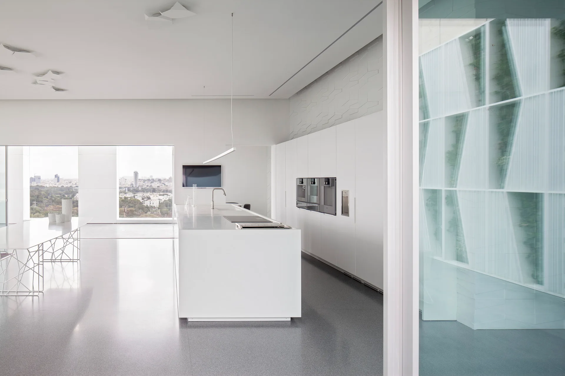 A minimalist white kitchen featuring a large central island, integrated stainless steel ovens, a grey floor, and a geometric wire base dining table.