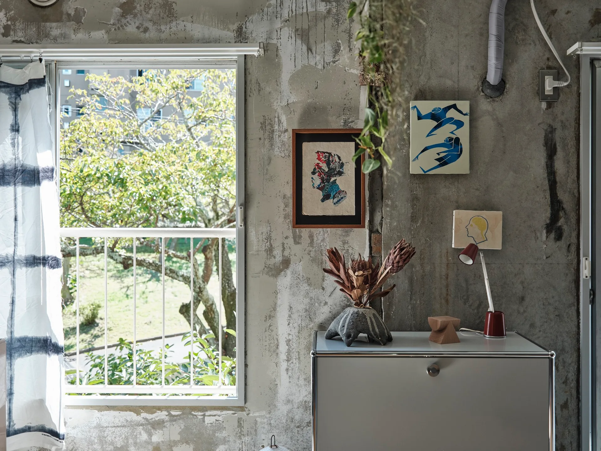 A sleek white modular cabinet sits against a raw concrete wall adorned with small art prints, next to a window showing a garden view.