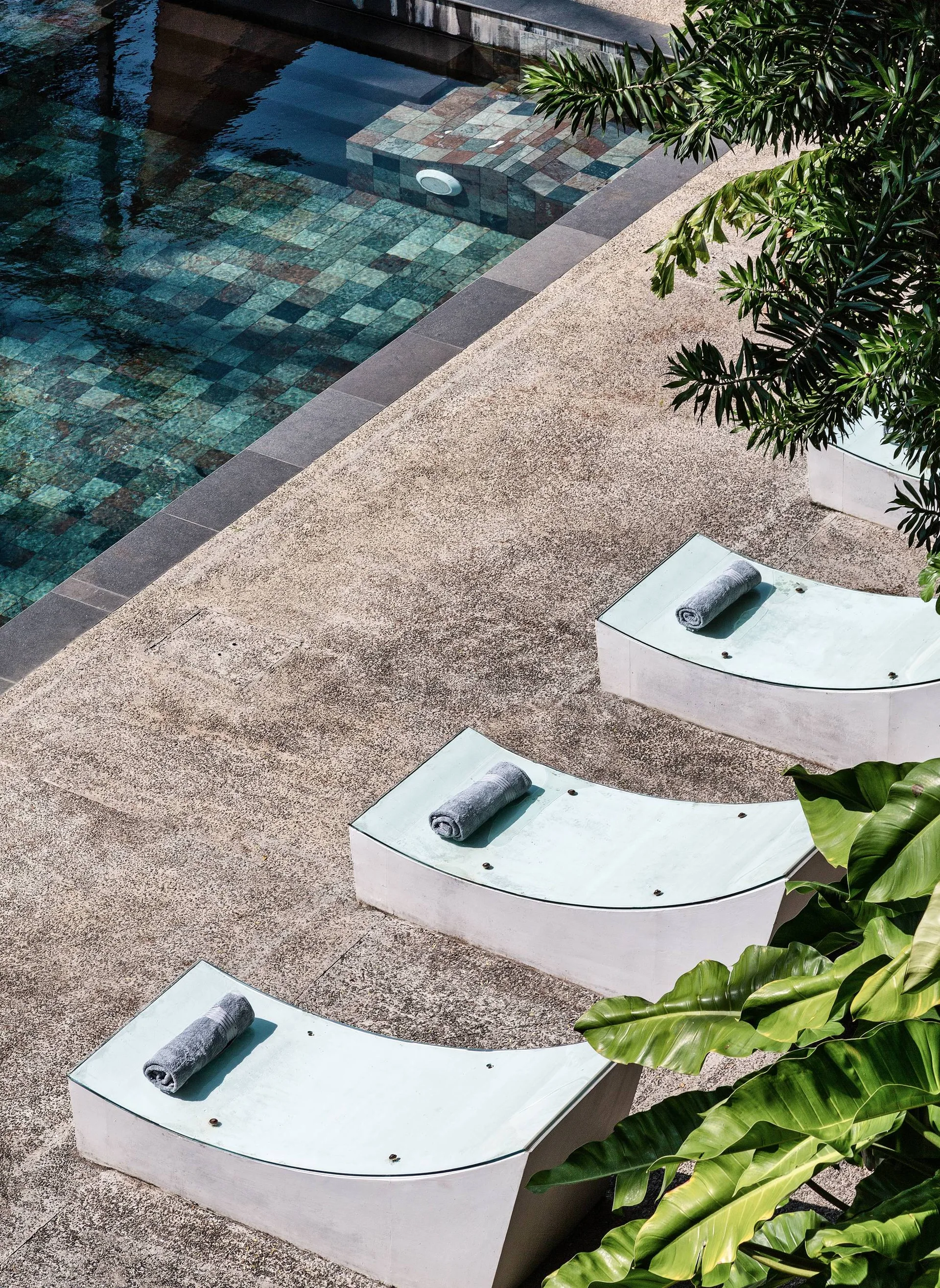 Three curved white lounge chairs with grey rolled towels sit on a textured tan deck beside a tiled swimming pool with tropical plants.