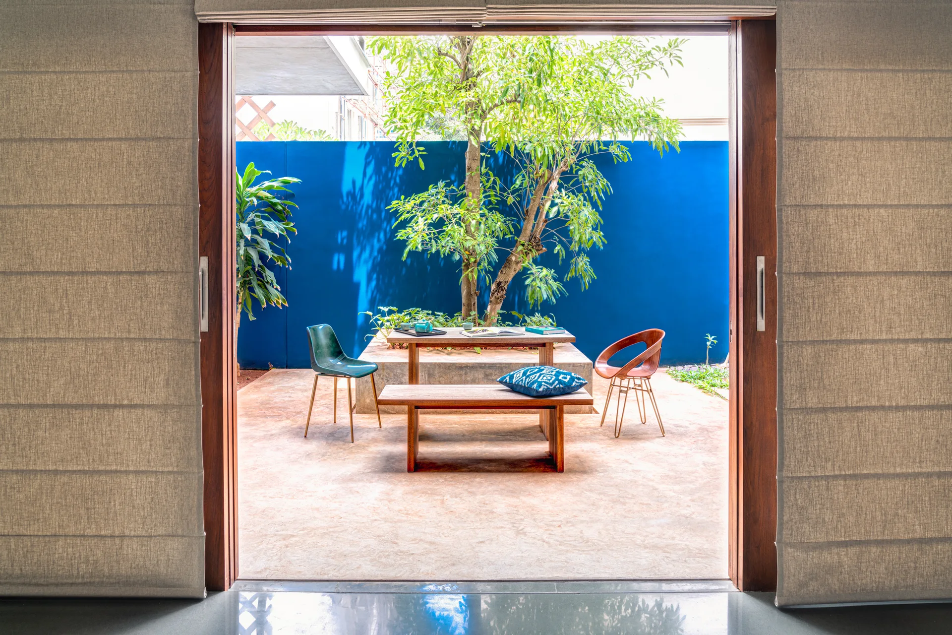 A wooden table and bench set with mid-century chairs sits in a sunlit courtyard framed by a sliding door and backed by a bright blue wall.