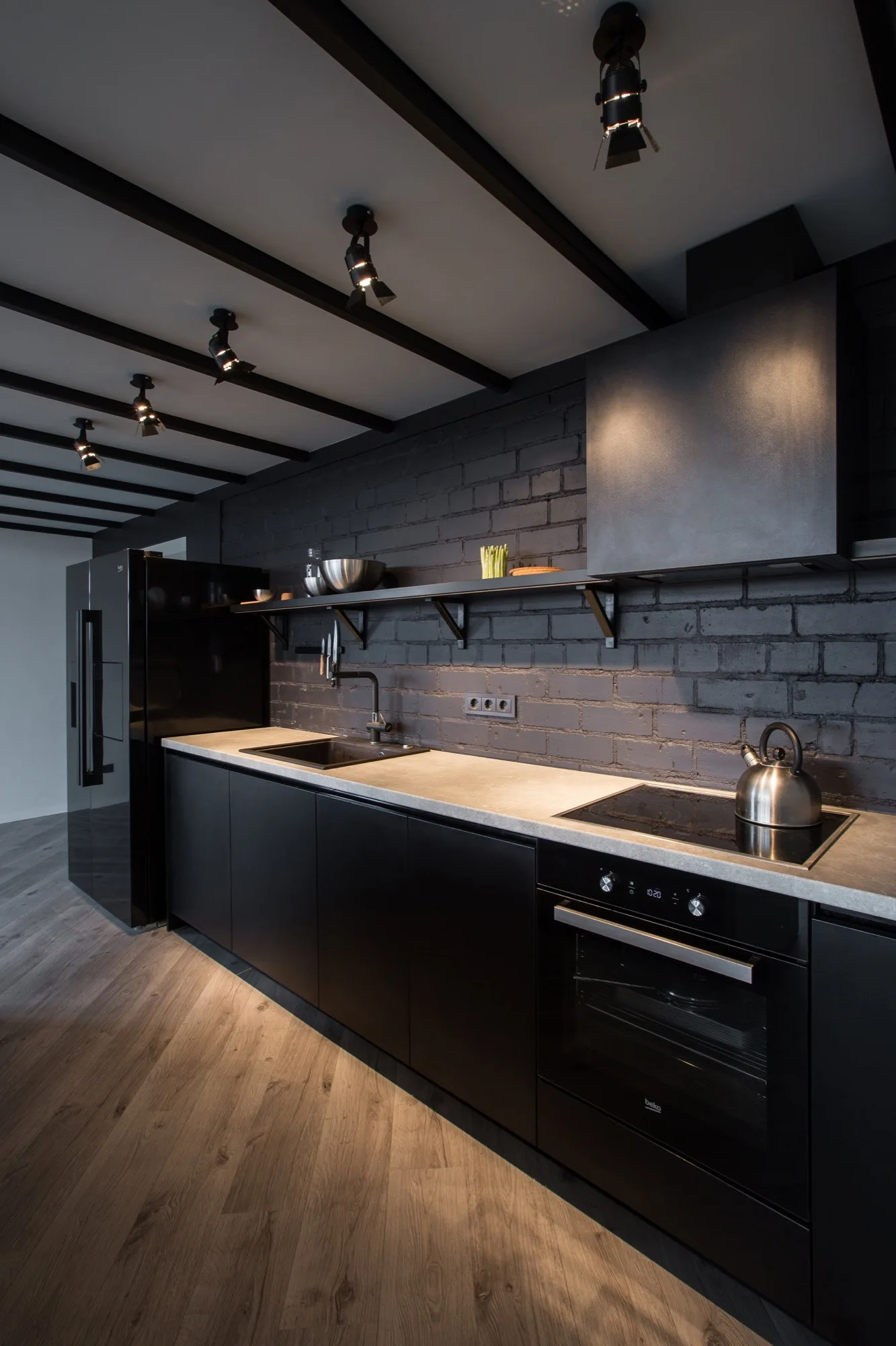 A modern industrial kitchen with matte black cabinetry, a dark painted brick wall, concrete countertops, and ceiling track lighting.