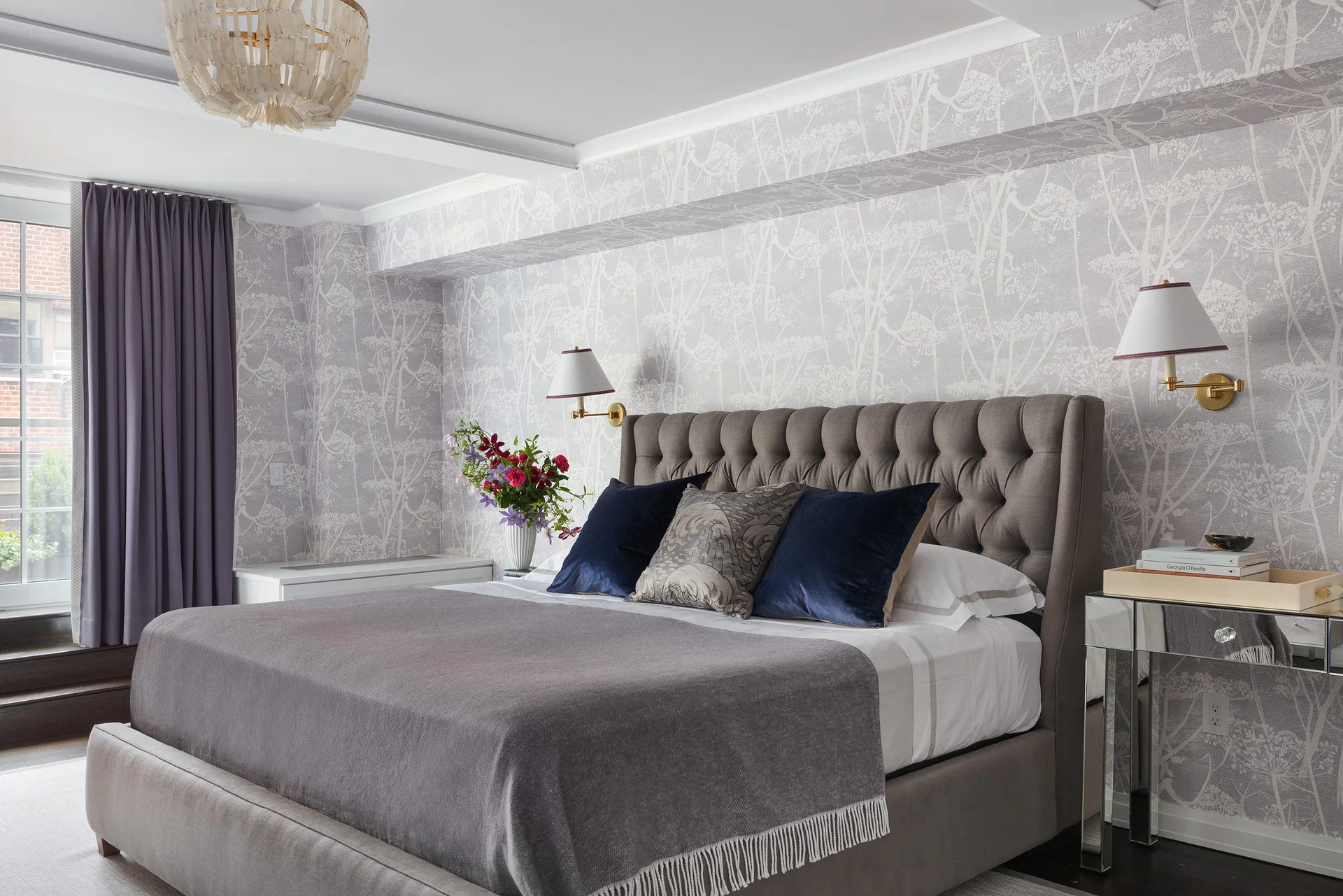 A bedroom features a grey tufted bed against botanical print wallpaper, flanked by a mirrored nightstand and purple curtains.