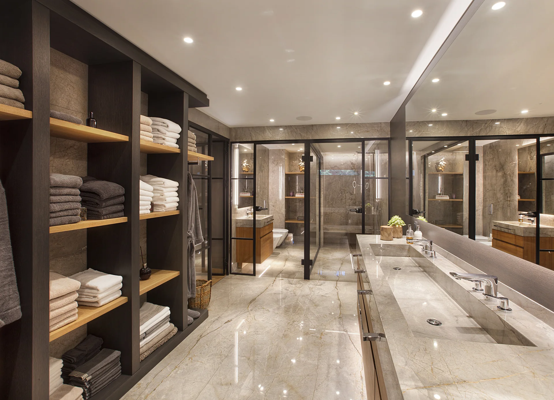 A spacious modern bathroom features a long marble double vanity, a large mirror, black metal glass doors, and a dark wood open shelving unit.