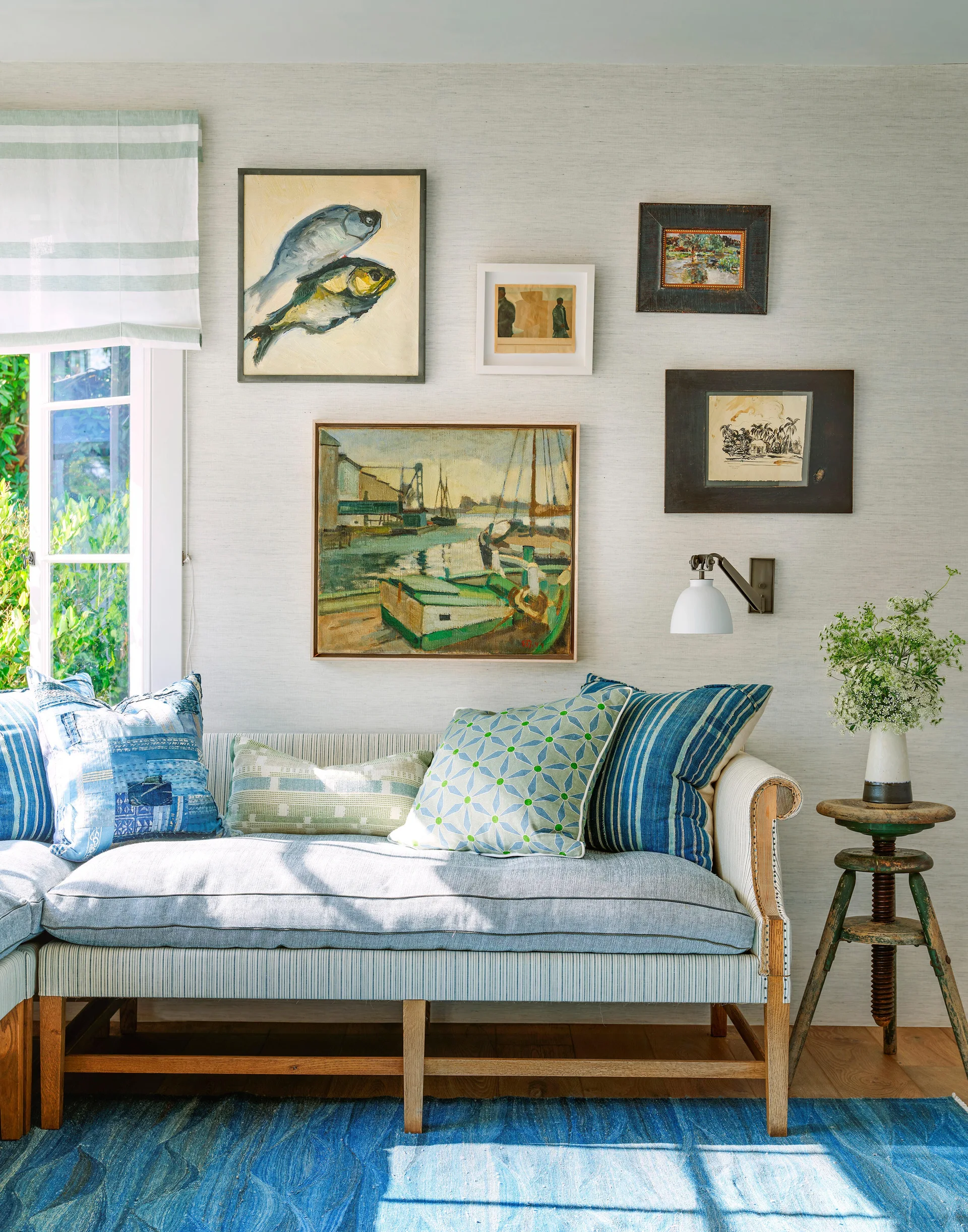 A blue striped upholstered bench sits against a grasscloth wall decorated with a gallery of framed art next to a vintage wooden stool.
