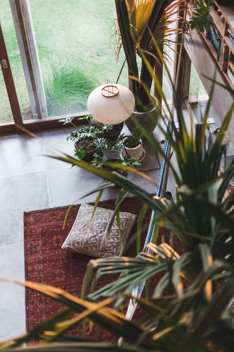A high-angle view of a grey-tiled living room with a red patterned rug, a geometric floor cushion, potted plants, and a white paper lantern.