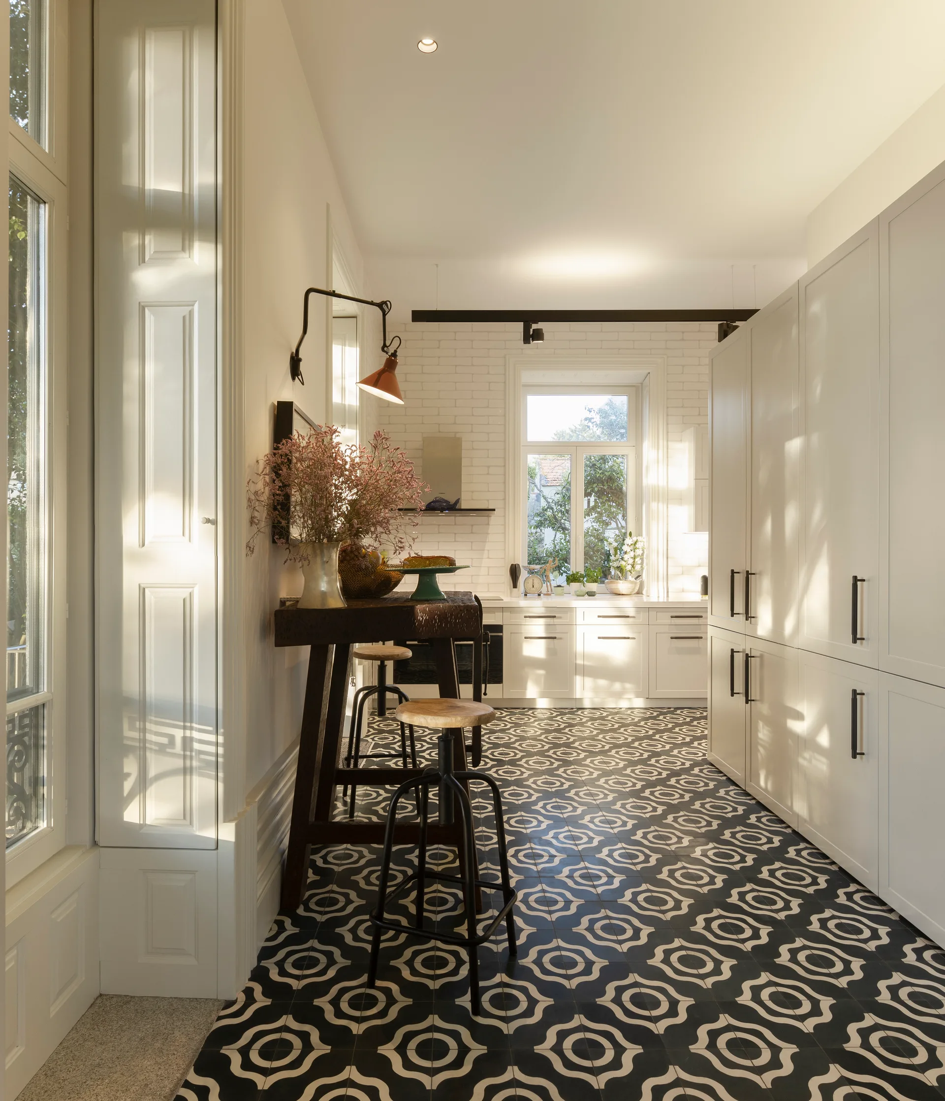 A bright galley kitchen features tall white cabinets, a narrow wooden breakfast bar with stools, and bold black and white geometric floor tiles.