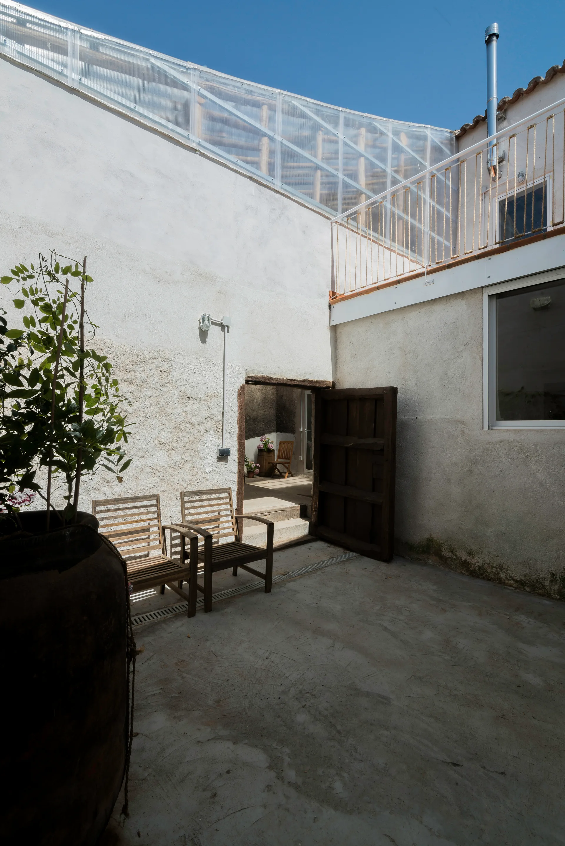 A rustic outdoor courtyard features white textured plaster walls, two wooden slat patio chairs, a large dark planter, and a balcony with rope railing.