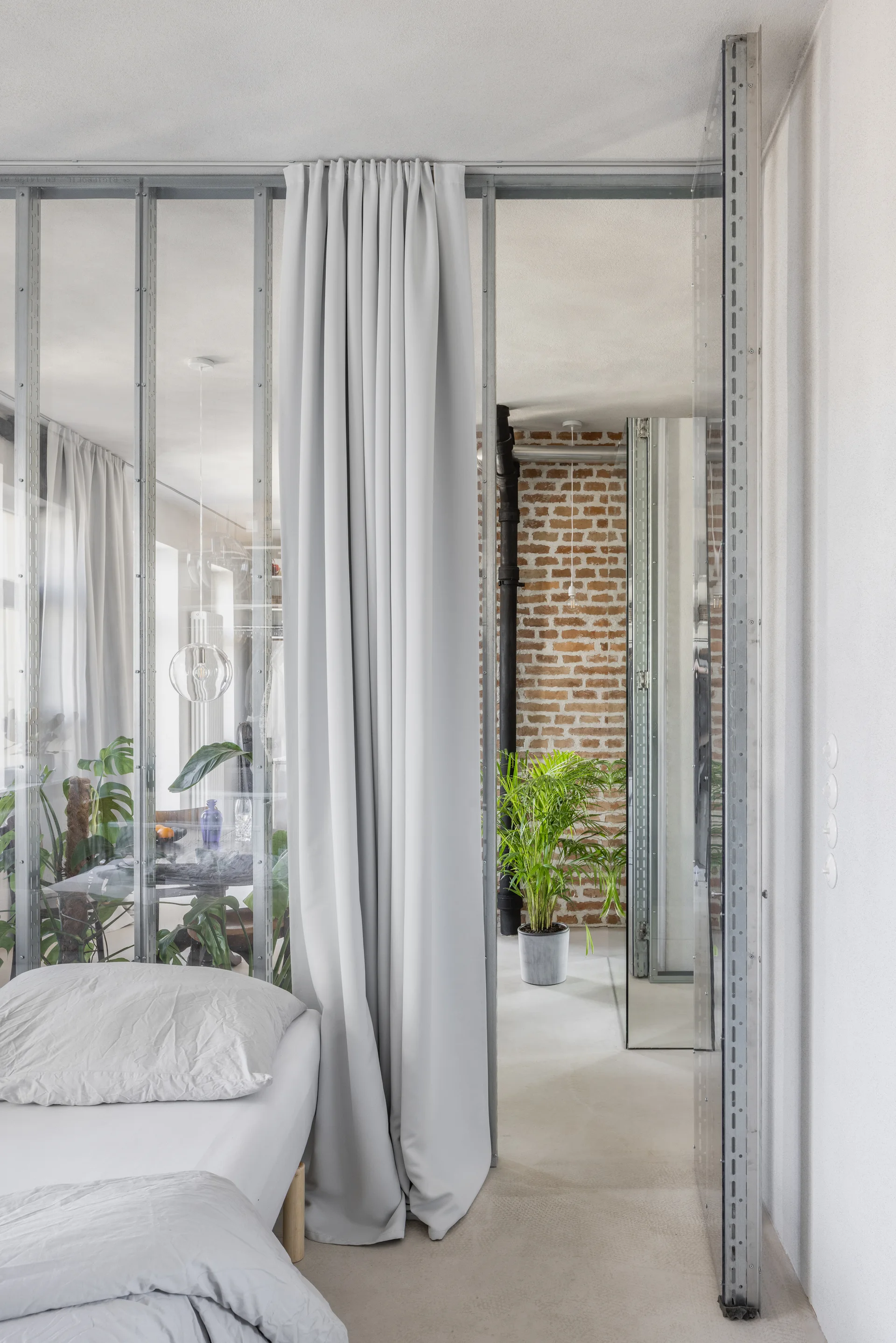 A bedroom features a light grey bed separated from an adjacent room by a metal and glass partition with a light grey floor-to-ceiling curtain.