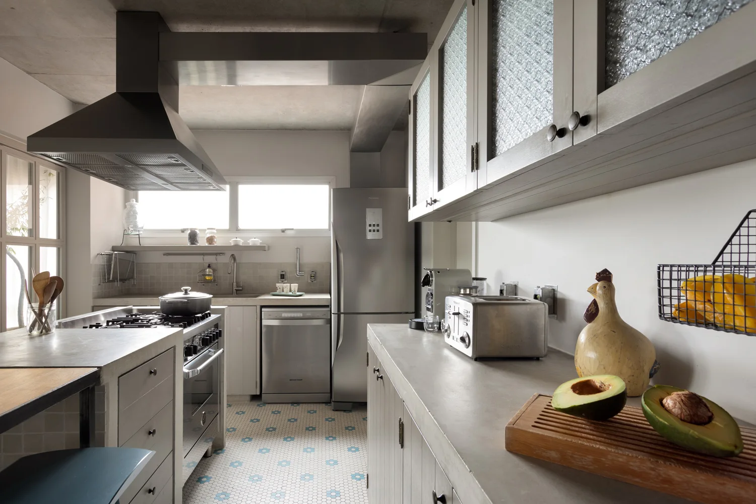 A modern kitchen featuring grey cabinets with textured glass doors, concrete countertops, a stainless steel gas range, and patterned hexagonal floor tiles.