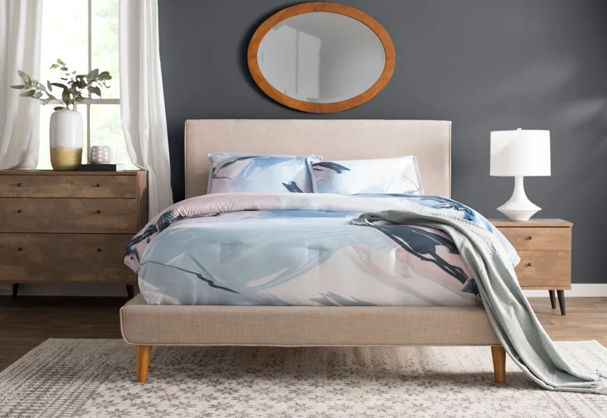 A beige upholstered bed with blue and pink abstract bedding is set against a dark gray wall with an oval wood mirror.