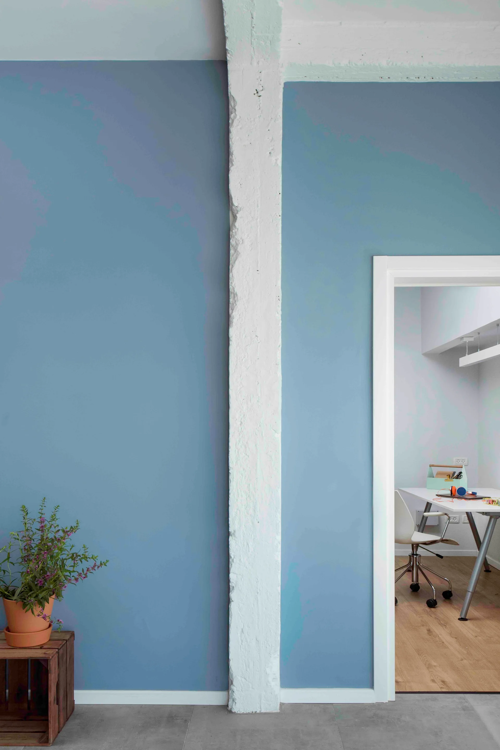 A textured white structural column separates a matte slate blue wall from a doorway revealing a bright home office with a desk and chair.