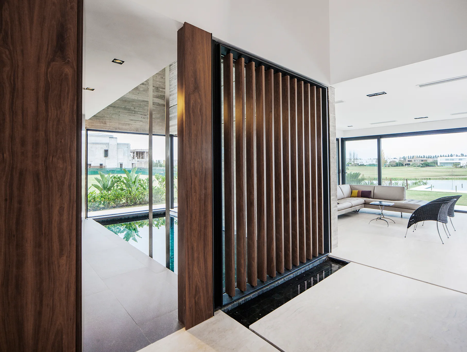 A modern living room features large floor tiles, a tan sectional sofa, a vertical wood slat screen, and an adjacent indoor water feature.