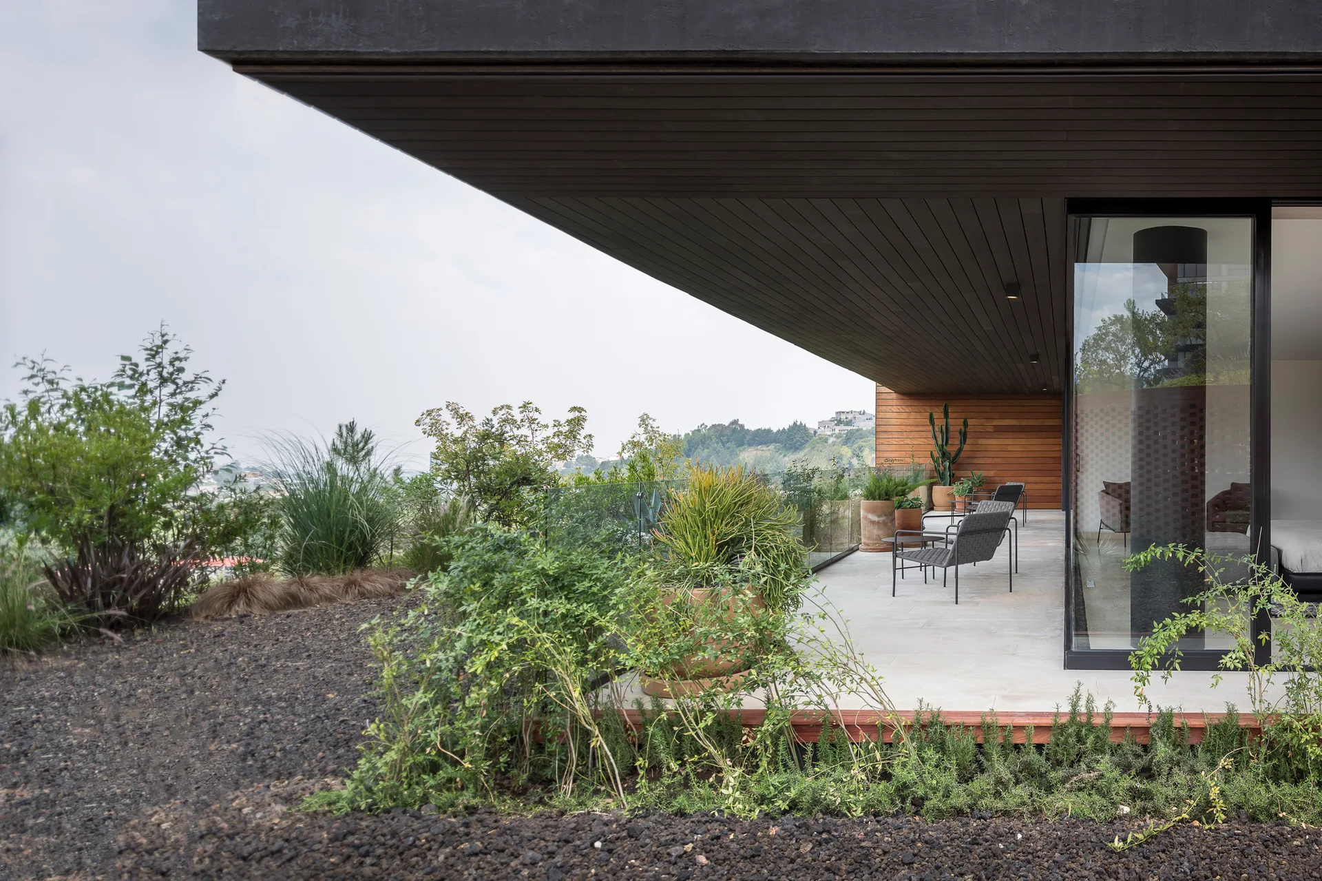 A modern covered terrace featuring a deep dark wood overhang, light stone tile flooring, modern patio chairs, and potted plants with a glass railing.