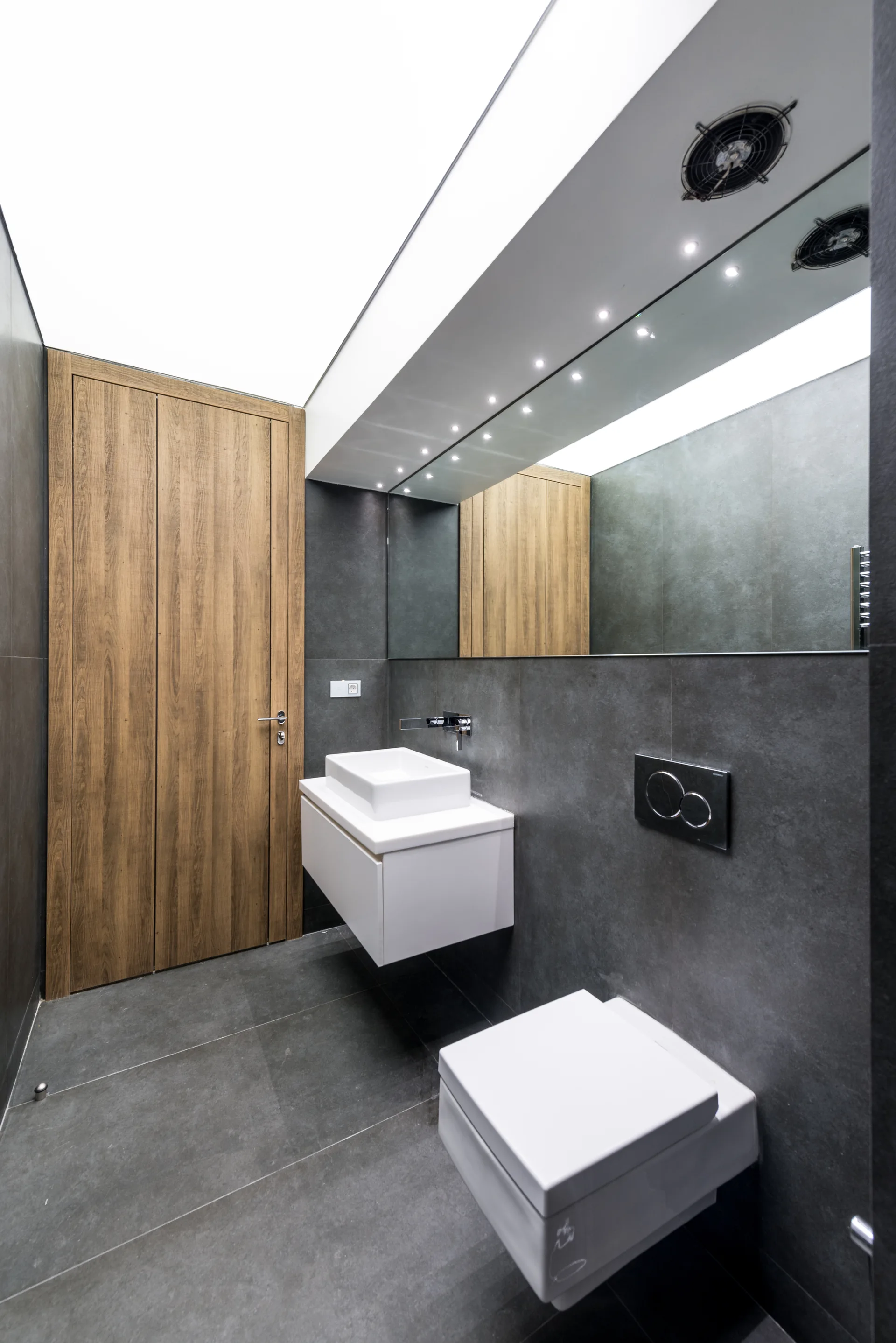 A contemporary bathroom featuring charcoal gray tiled walls, a floating white vanity with a rectangular sink, a wall-hung toilet, and a wood grain door.