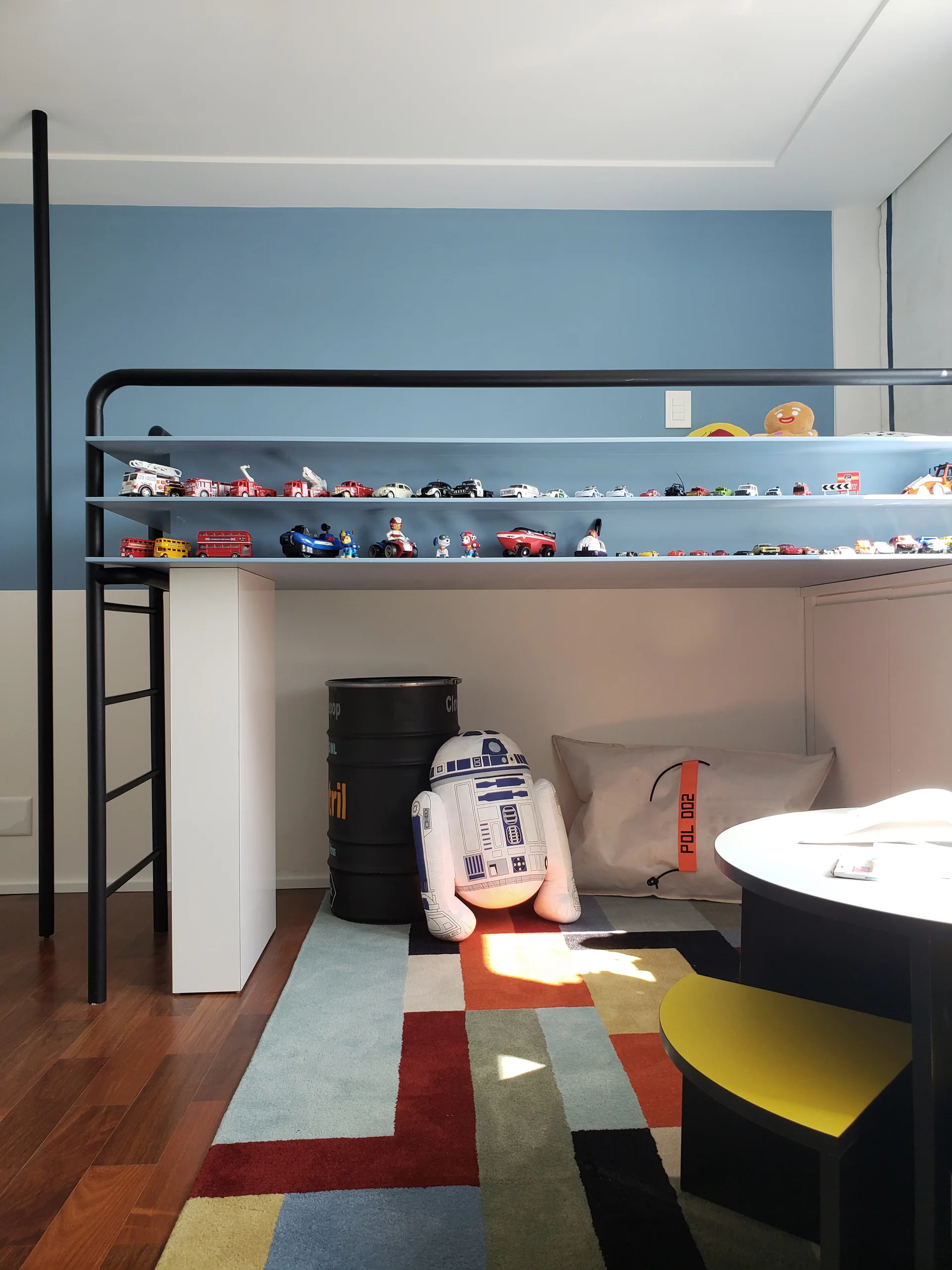 A children's bedroom features a black metal loft bed with blue built-in shelves displaying toy cars above a colorful geometric rug.