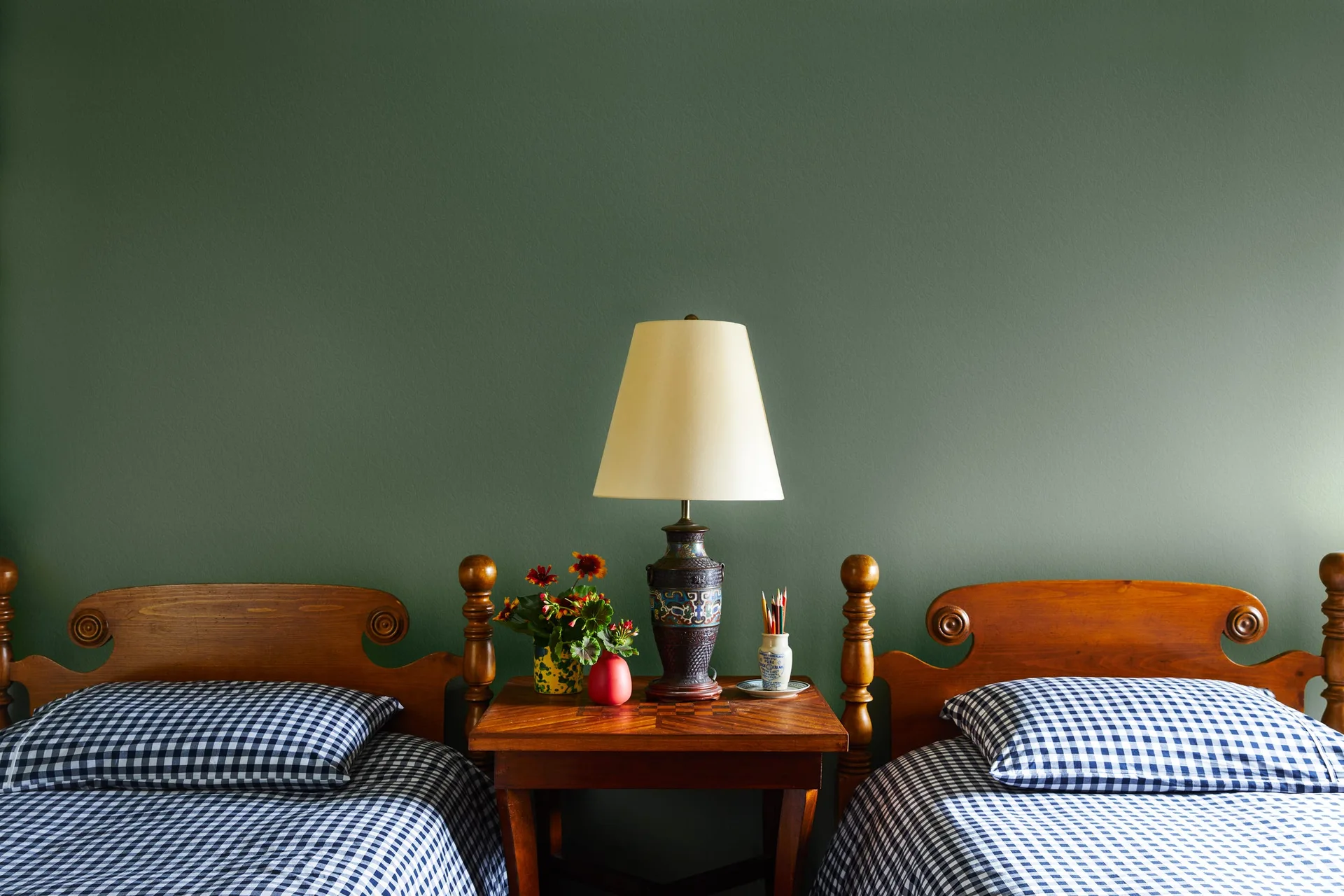 Two matching wooden twin beds with blue gingham bedding flank a wood nightstand with a ceramic lamp against a solid green wall.