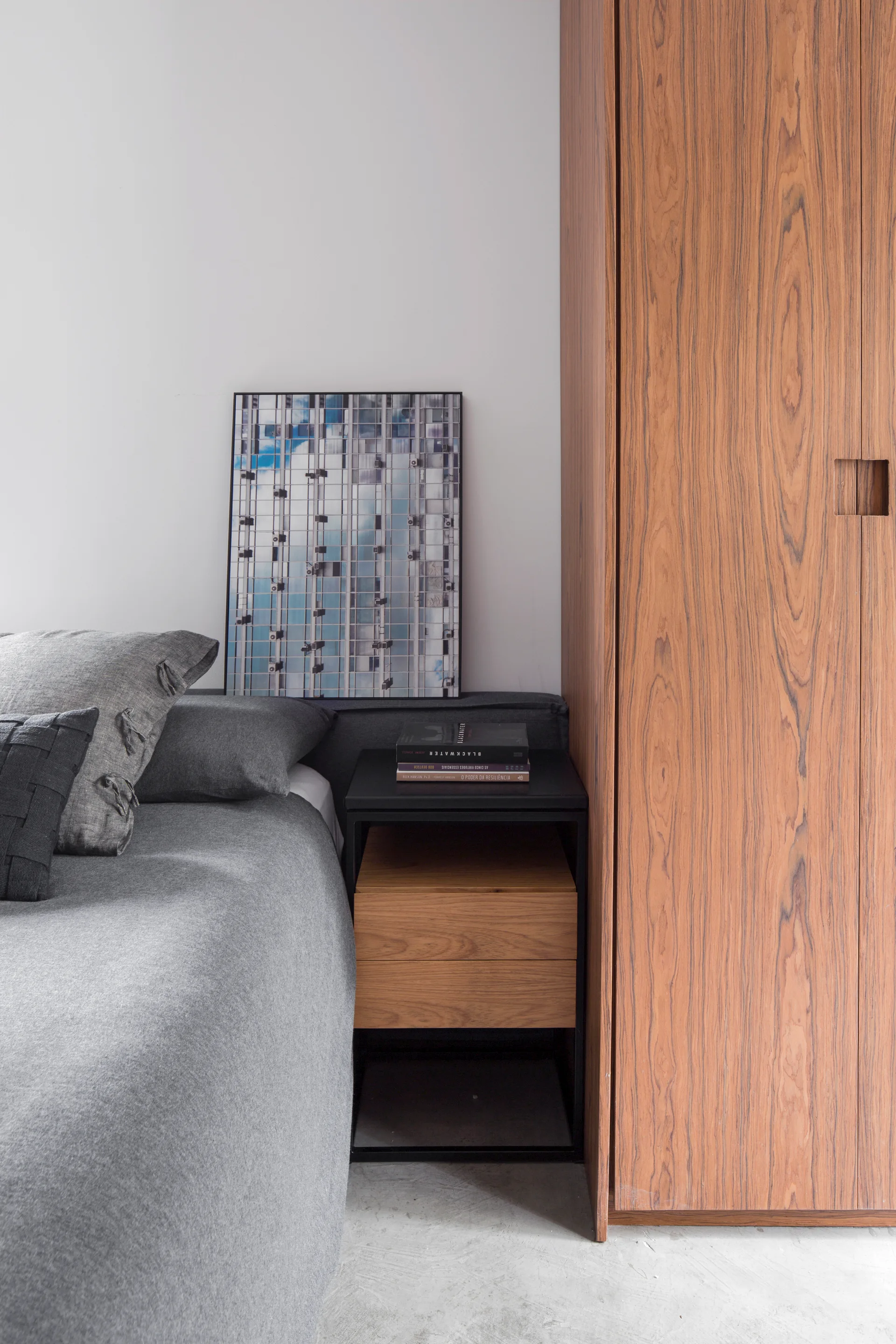 A minimalist bedroom featuring a grey bed, a black-framed nightstand, and a tall wood grain wardrobe.