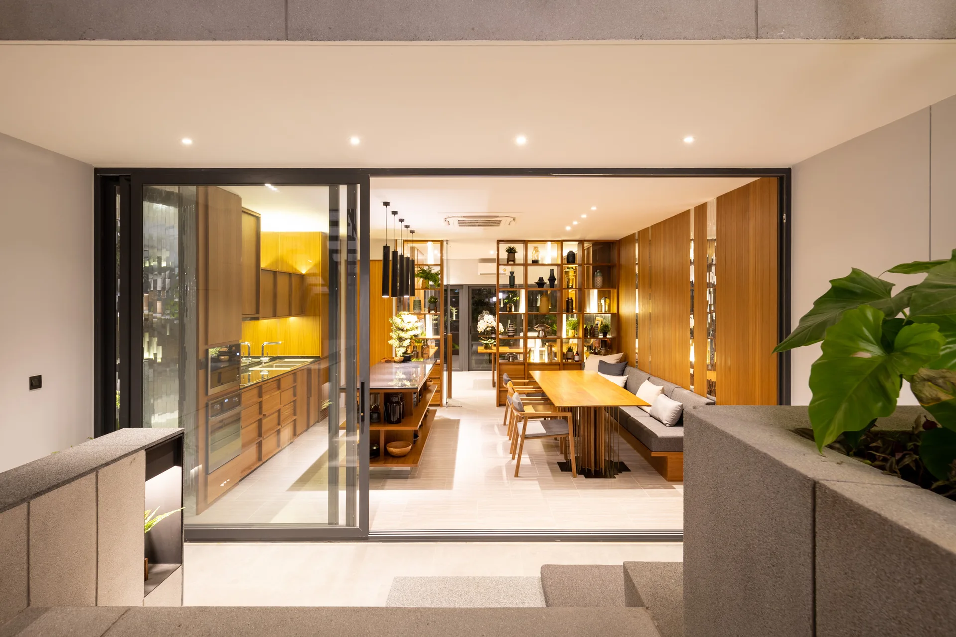 A modern open-plan kitchen and dining area with warm wood cabinetry, a built-in bench, and a dining table viewed through a sliding glass door.