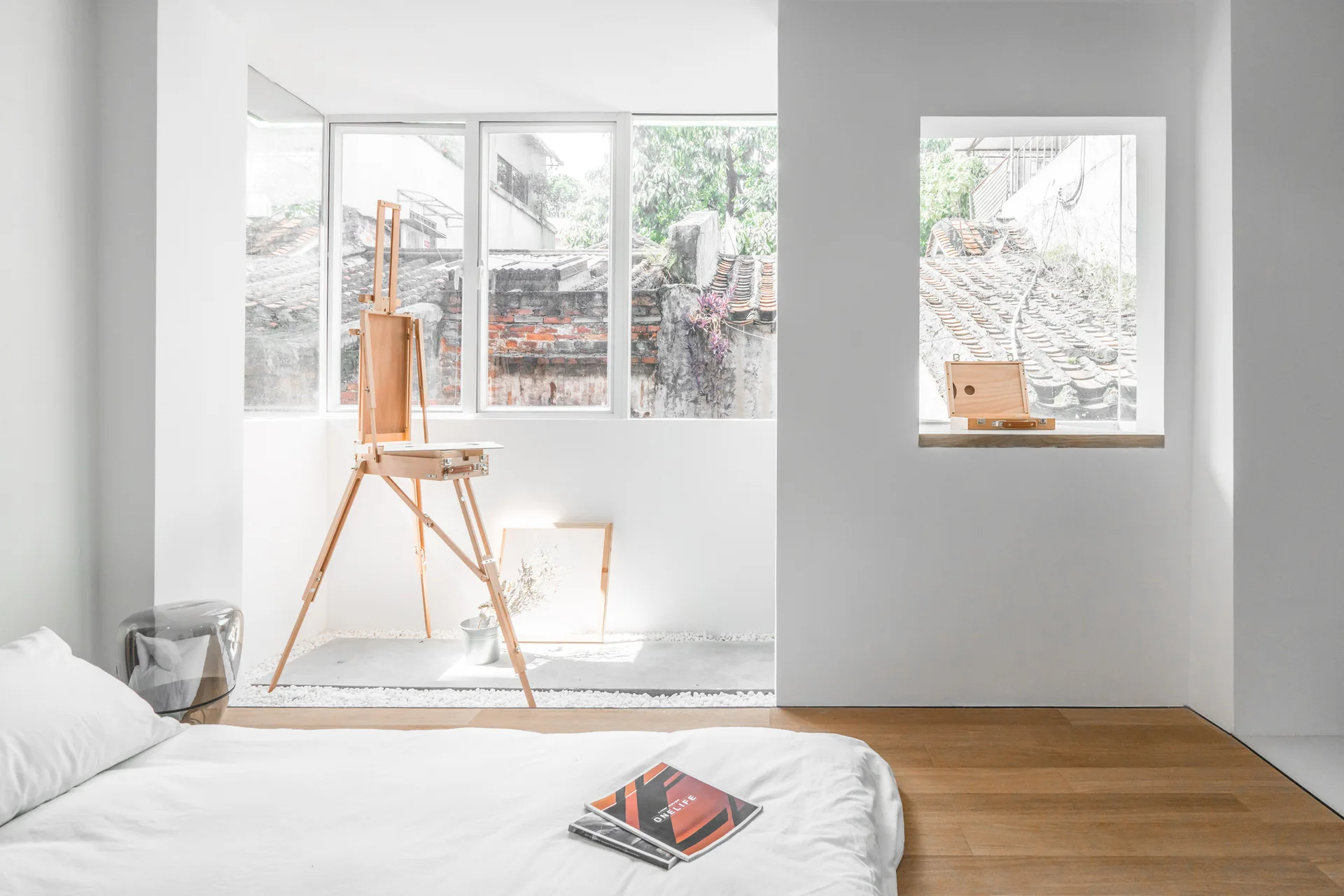 A minimalist bedroom features a low bed on wood floors and a wooden artist easel standing in a pebble-filled window niche.