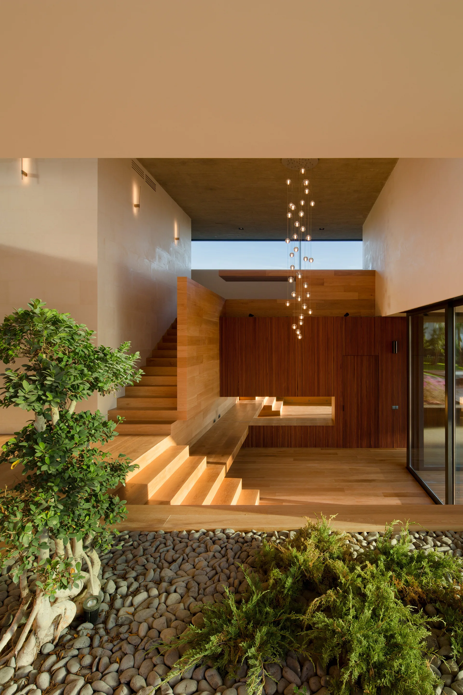 A modern wood staircase rises past a slatted timber wall and a multi-globe pendant light, with an indoor pebble garden in the foreground.