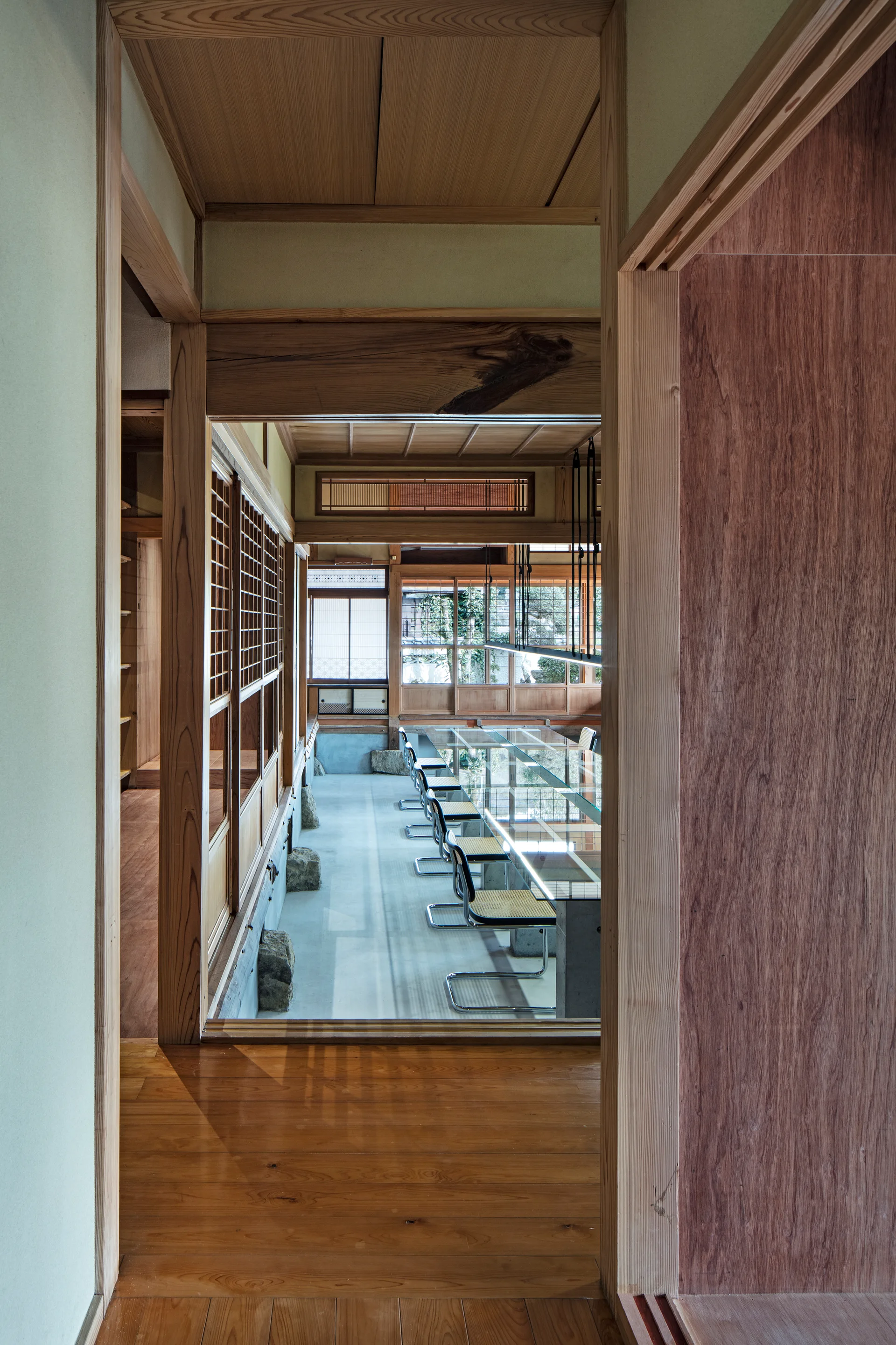 A view through a wooden doorway into a Japanese modern office featuring a long glass table, woven chairs, and wooden lattice sliding doors.