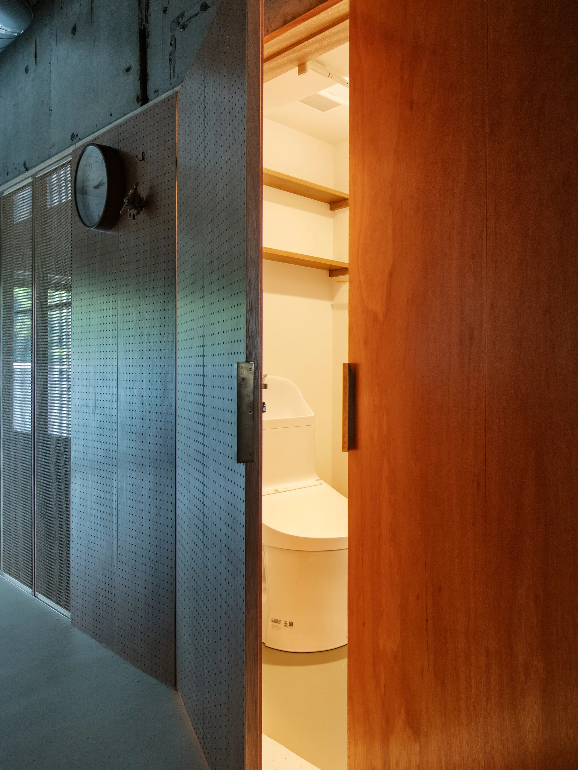 A wooden sliding door is partially open to reveal a minimalist bathroom with a white toilet and shelves under an exposed concrete ceiling.