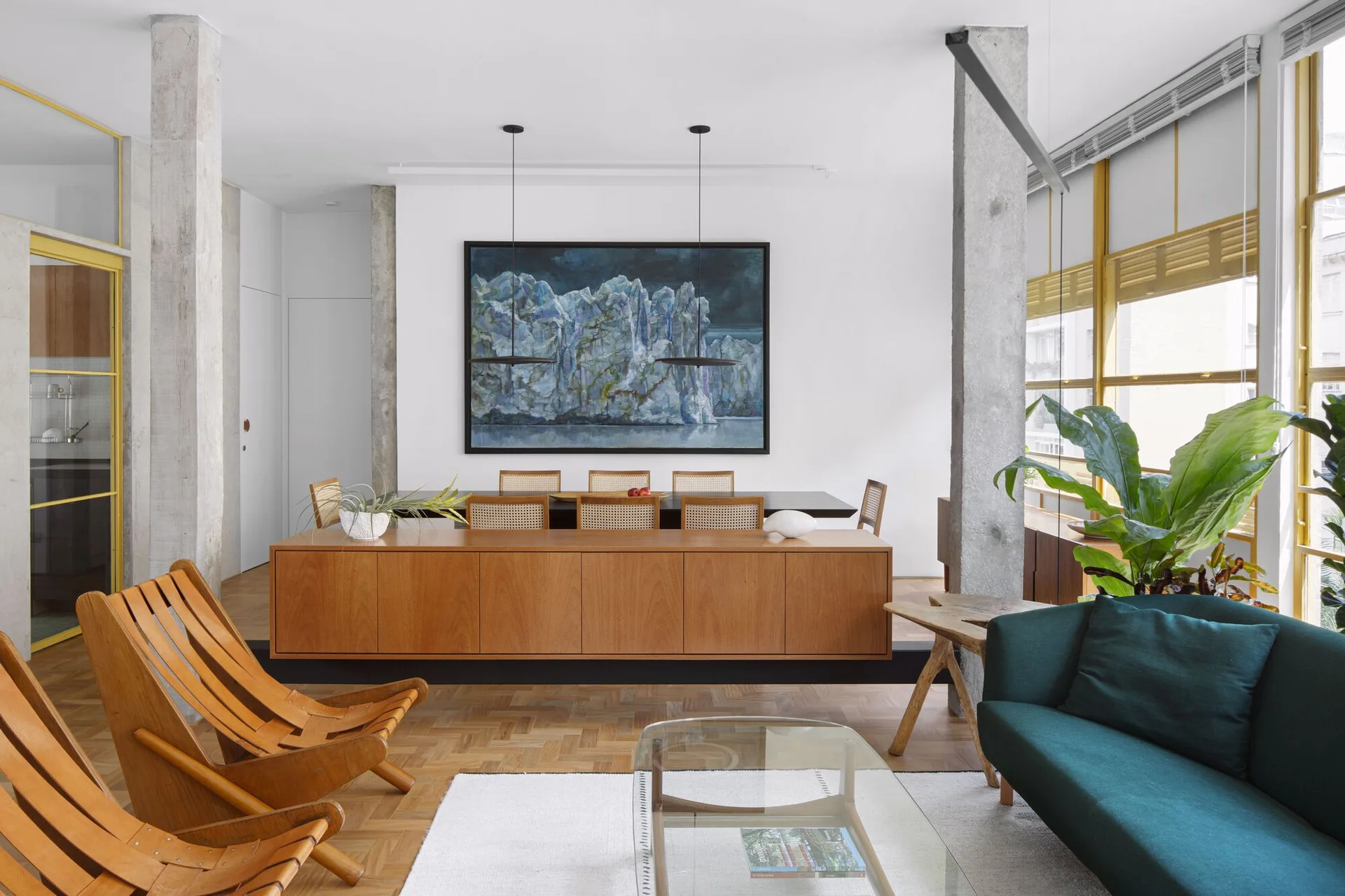 An open-concept living and dining area features exposed concrete columns, a long wooden sideboard, and a dark green sofa on herringbone wood floors.
