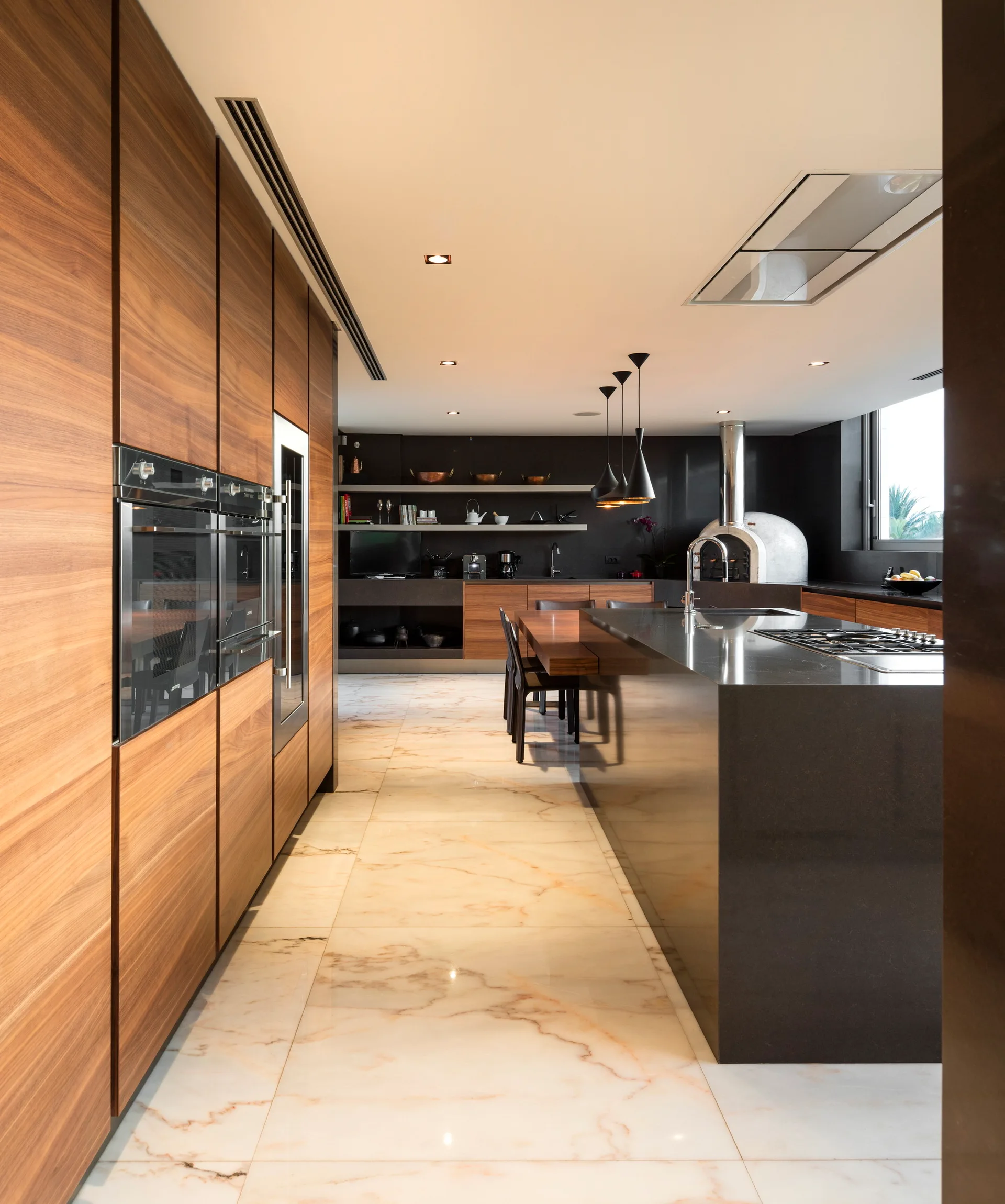 A modern kitchen featuring seamless wood-toned wall cabinets, a large dark stone island, and polished marble flooring with reddish-brown veining.