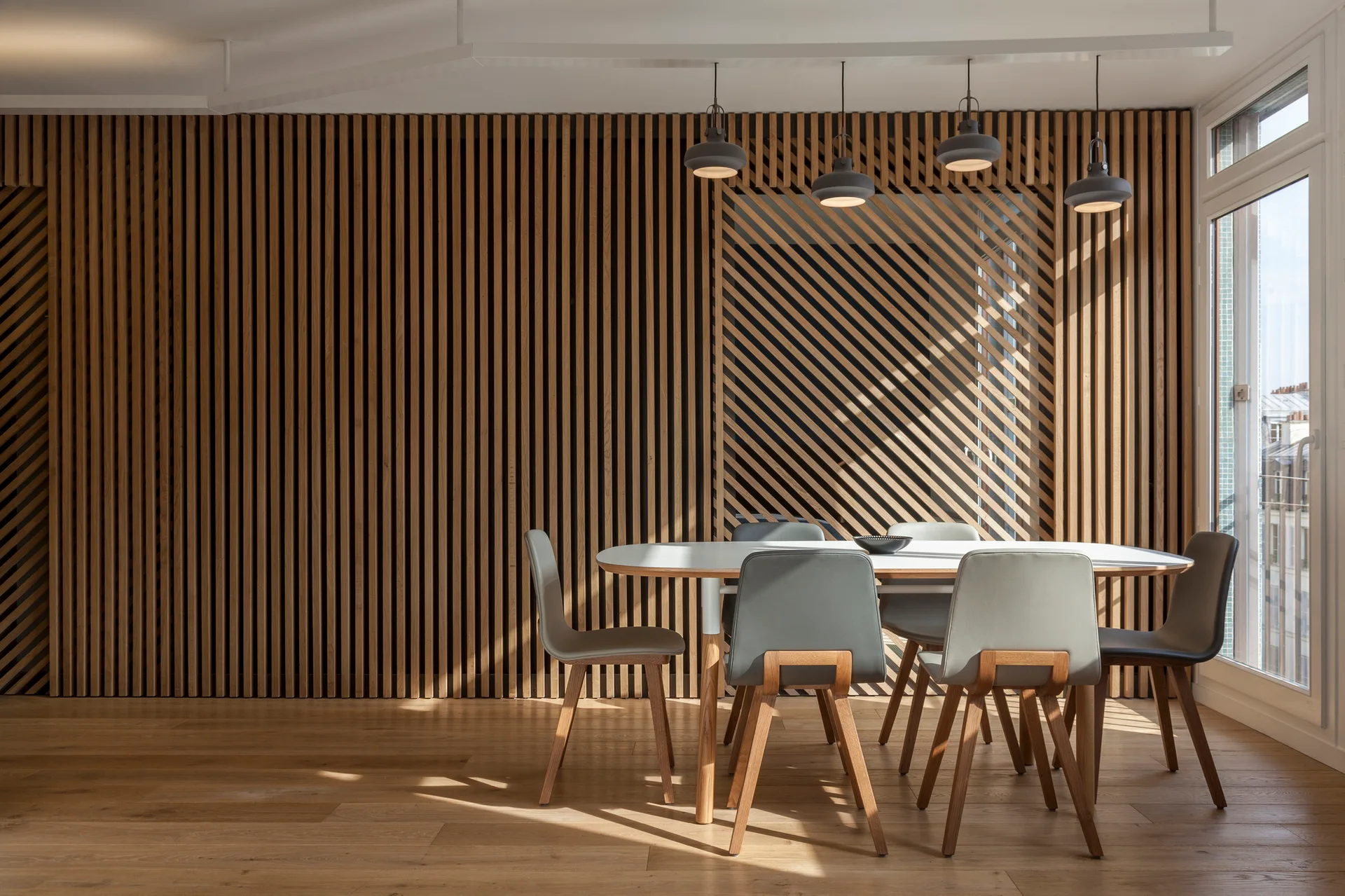 A modern dining room features an oval wooden table surrounded by grey chairs set against a full-height vertical wood slat accent wall.