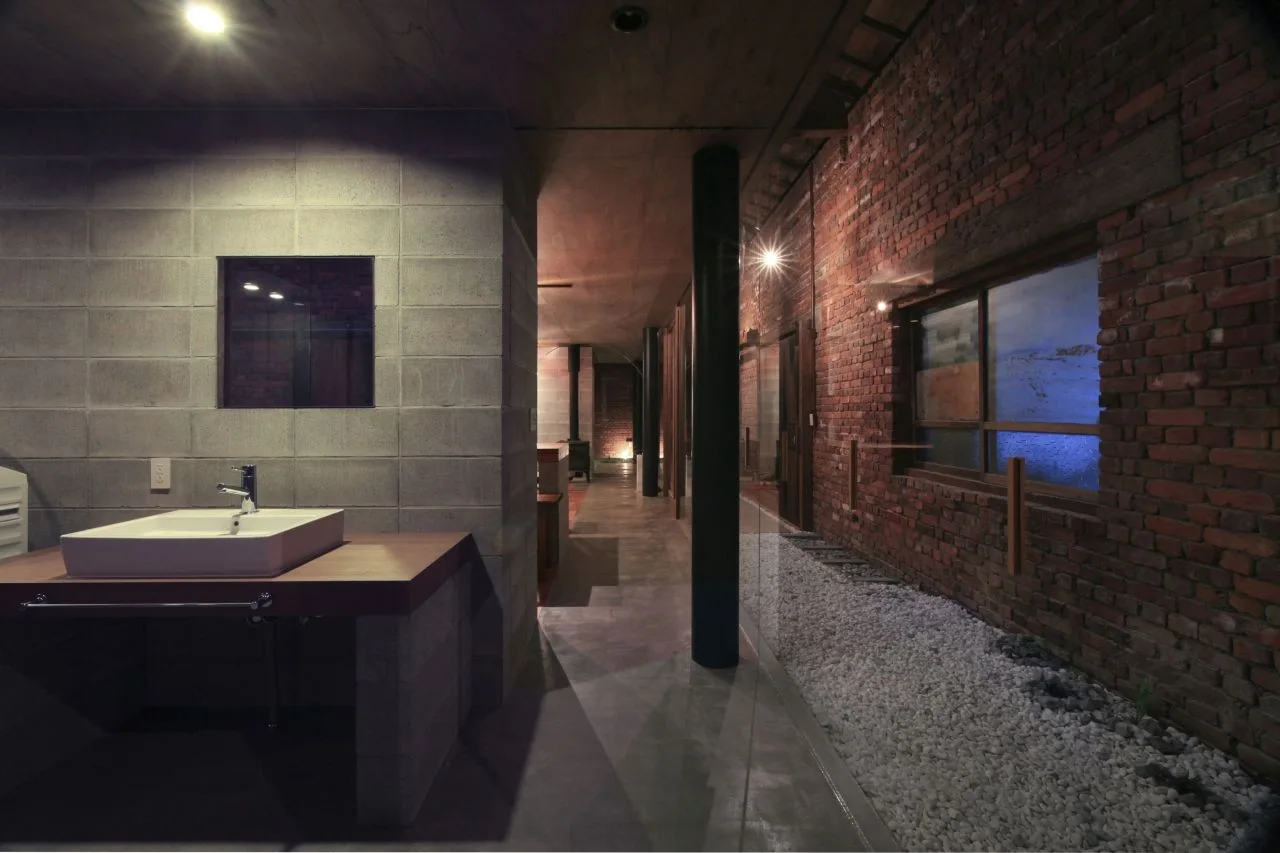 An industrial bathroom features a white vessel sink on a wooden countertop supported by concrete block, across from an exposed red brick wall.