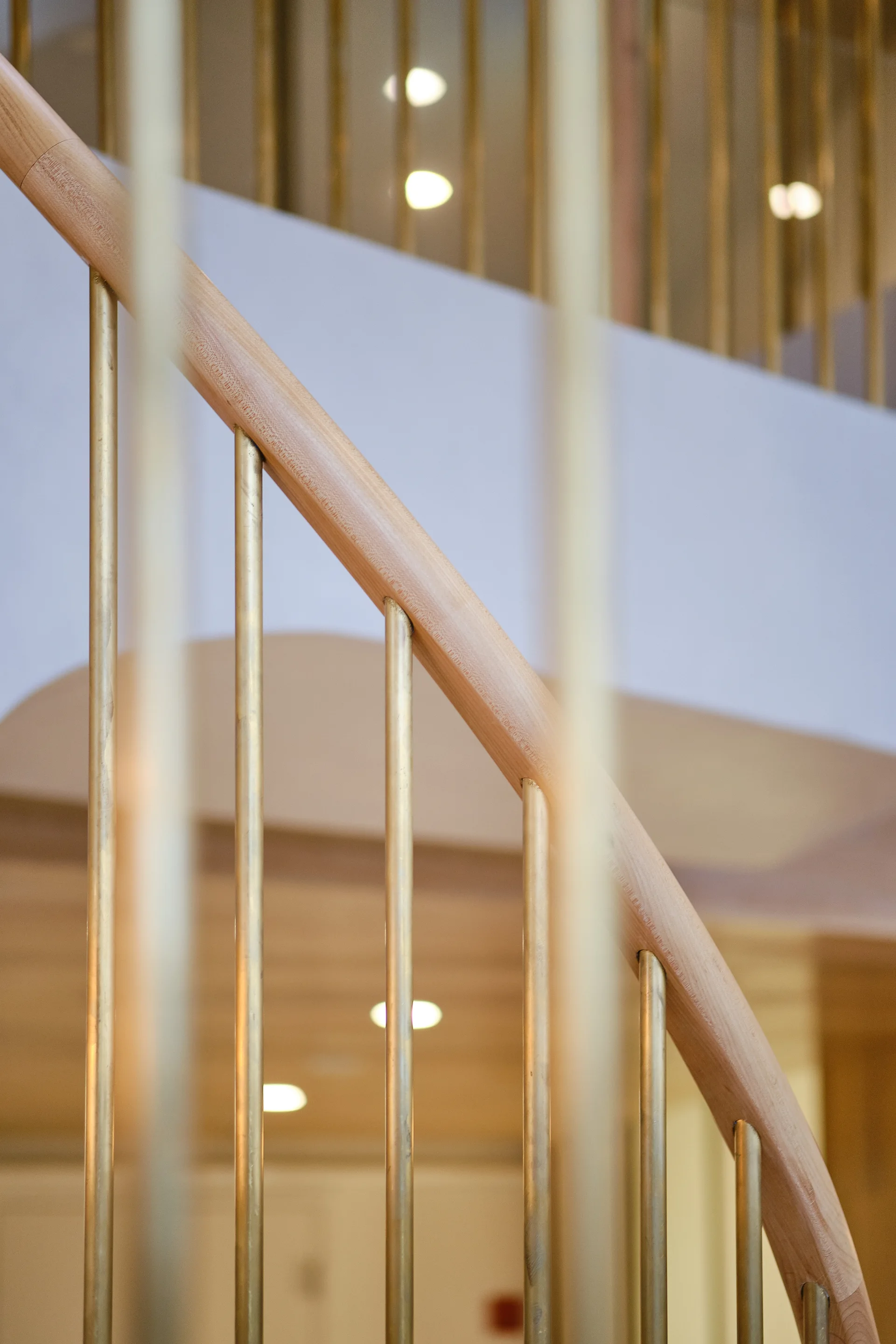 A close-up of a curved natural wood handrail with vertical brass-toned metal balusters on a modern staircase.