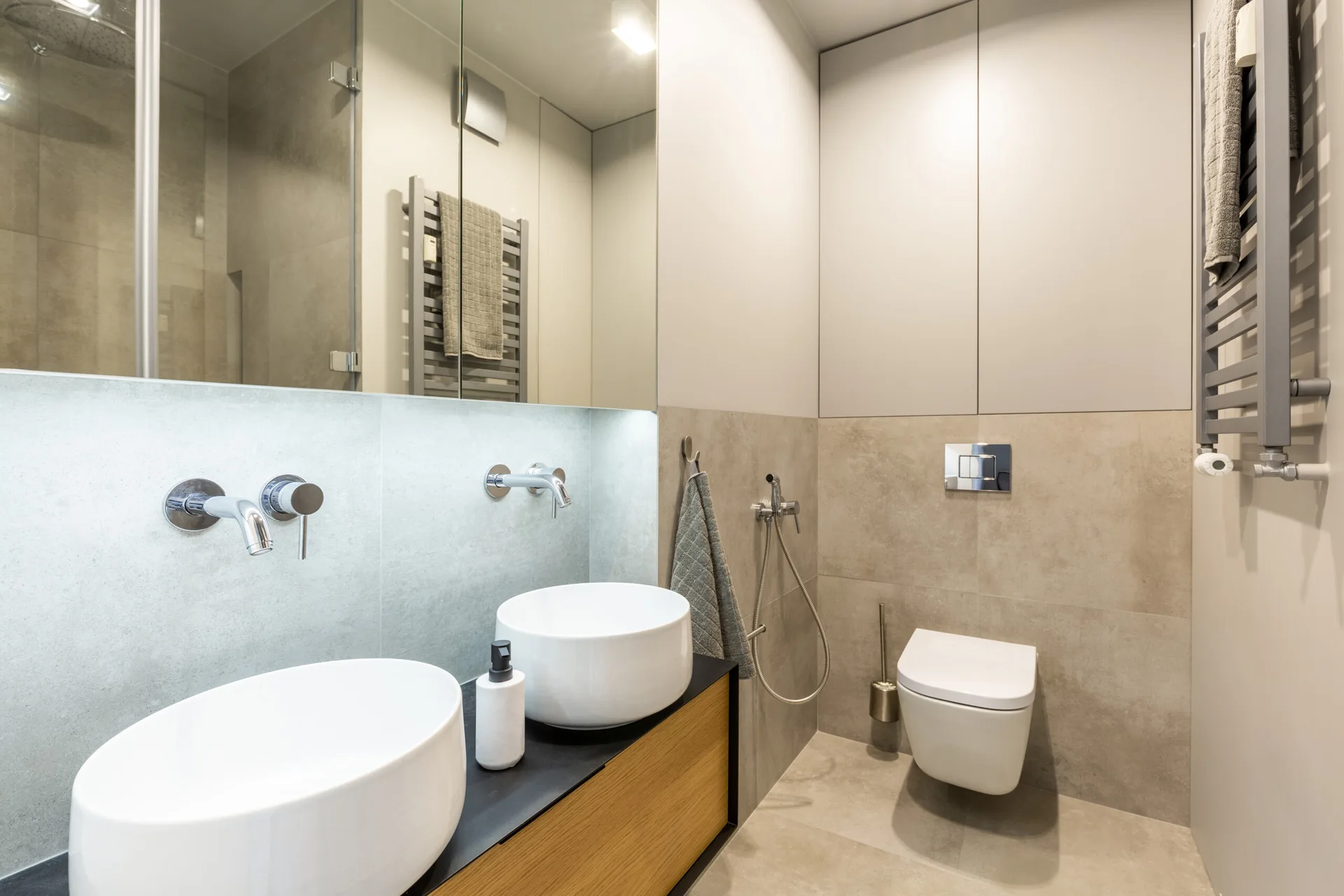 A contemporary bathroom features a wood-front vanity with two white round vessel sinks, wall-mounted faucets, a large mirror, and a wall-mounted toilet.