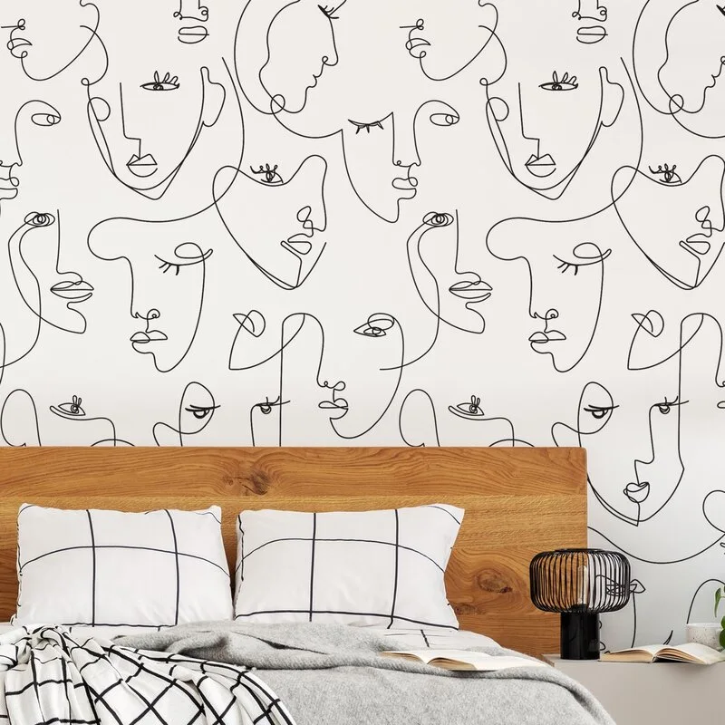 A bedroom features a natural wood headboard set against black and white line-art face wallpaper, accented by grid-patterned pillows.