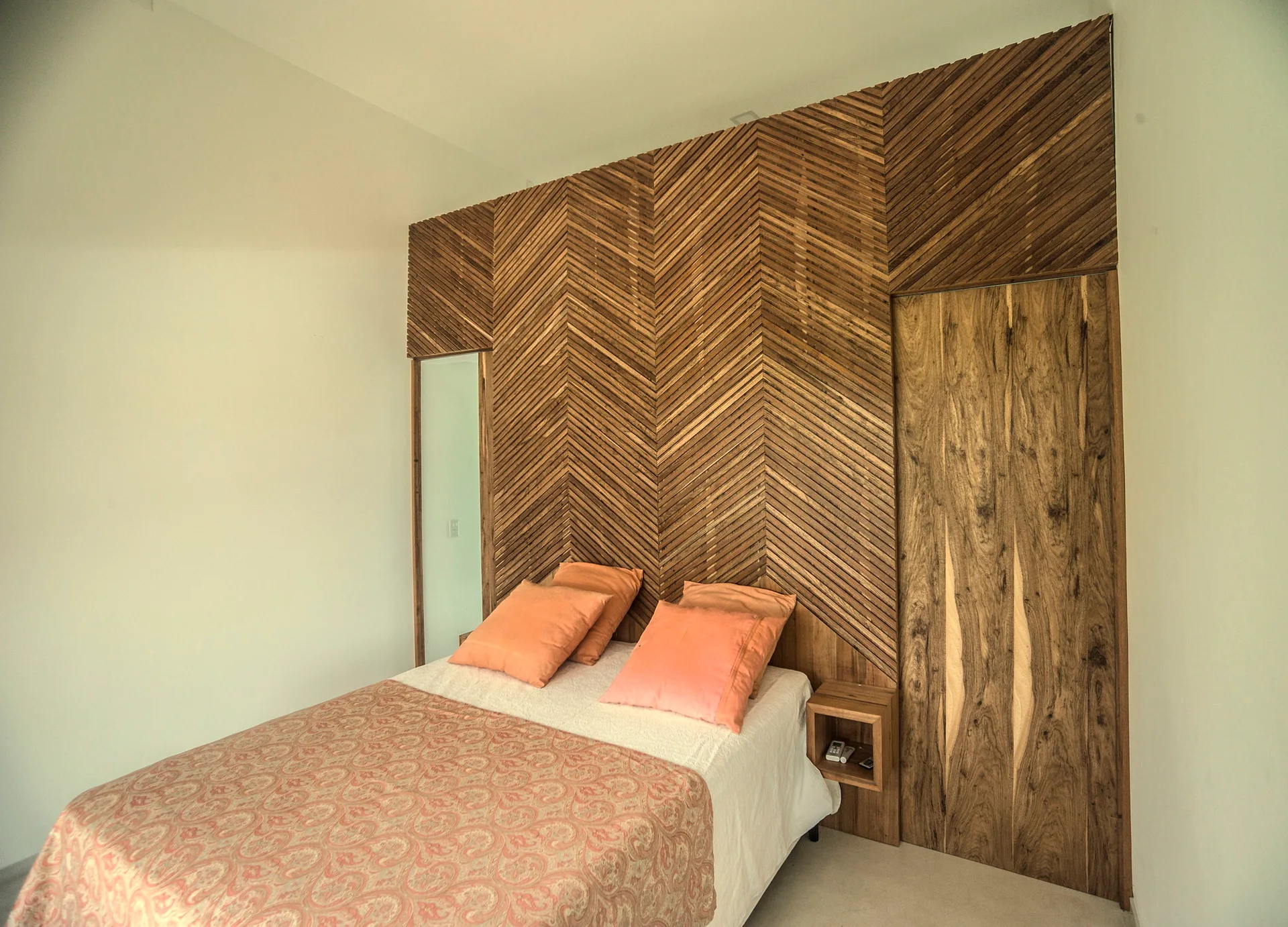 A bedroom features a bed with peach pillows set against a tall herringbone wood slat wall with an integrated bedside cubby and a wood door.