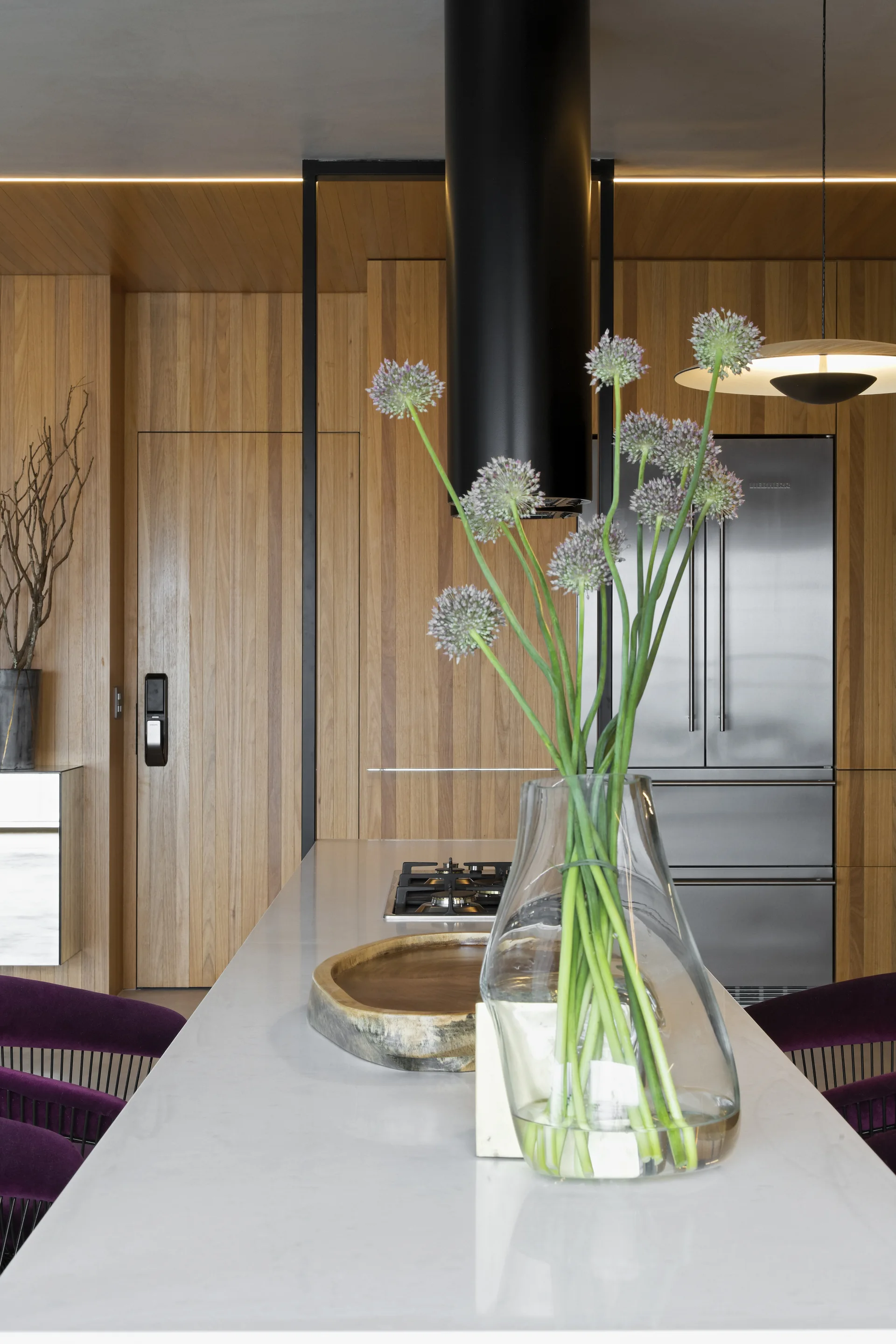 A modern kitchen island with a light countertop and purple velvet stools sits before a wall of vertical wood paneling with integrated appliances.