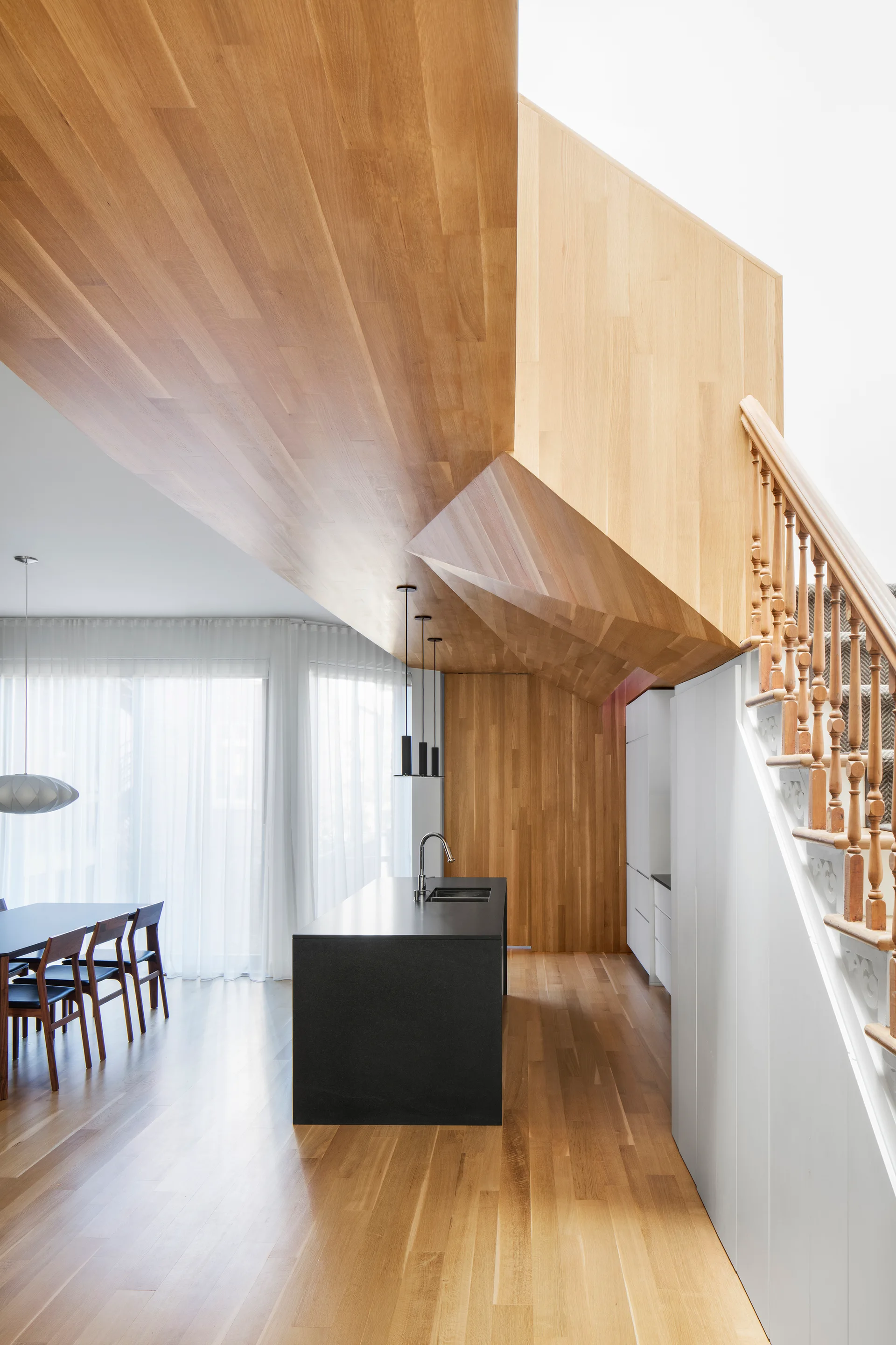A modern kitchen features a black island under a sculpted wood-paneled ceiling, with a wooden staircase and dining area nearby.