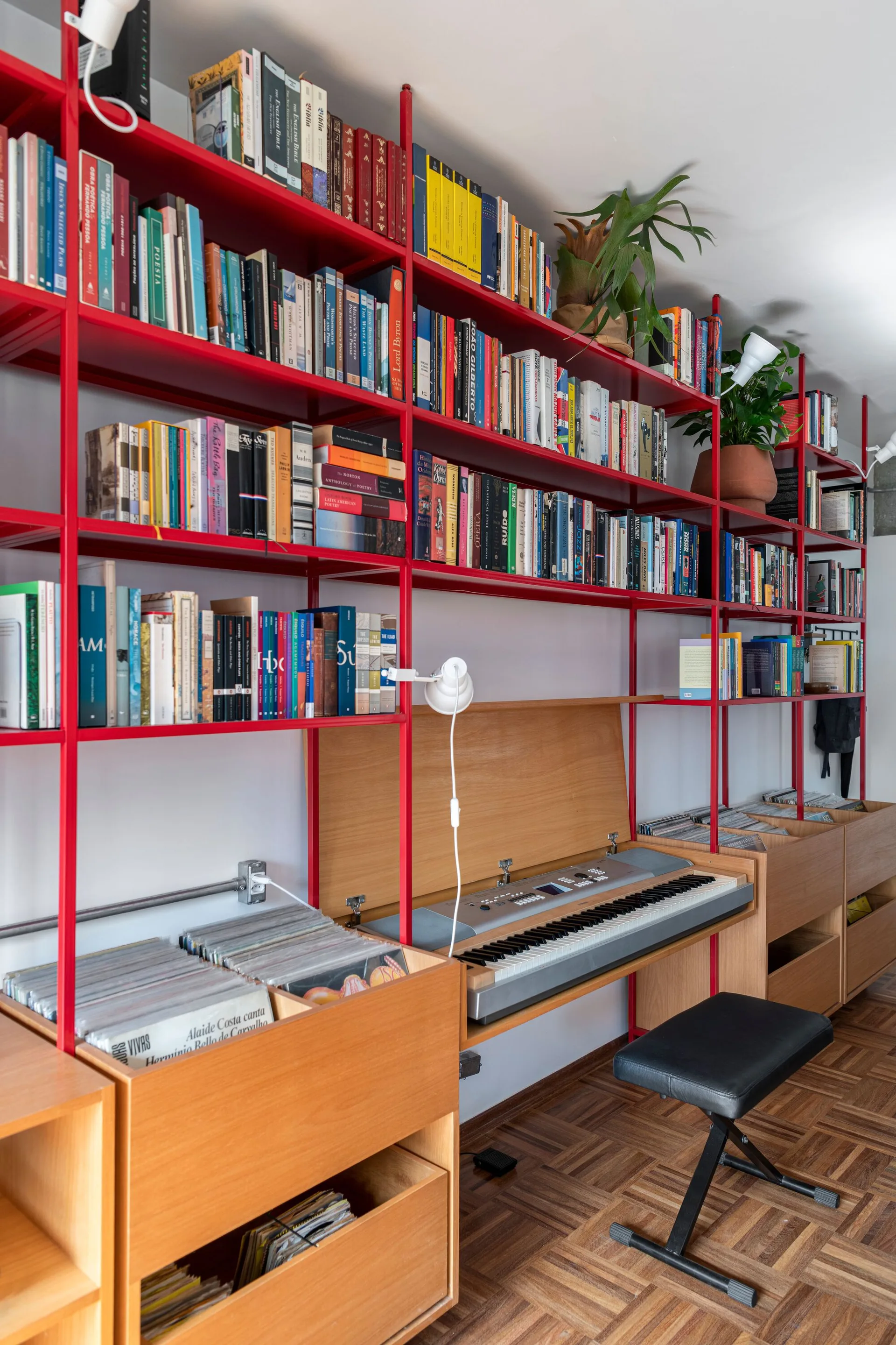 A vibrant red metal modular shelving unit holding books with integrated wooden vinyl record storage bins, a keyboard, and task lights.