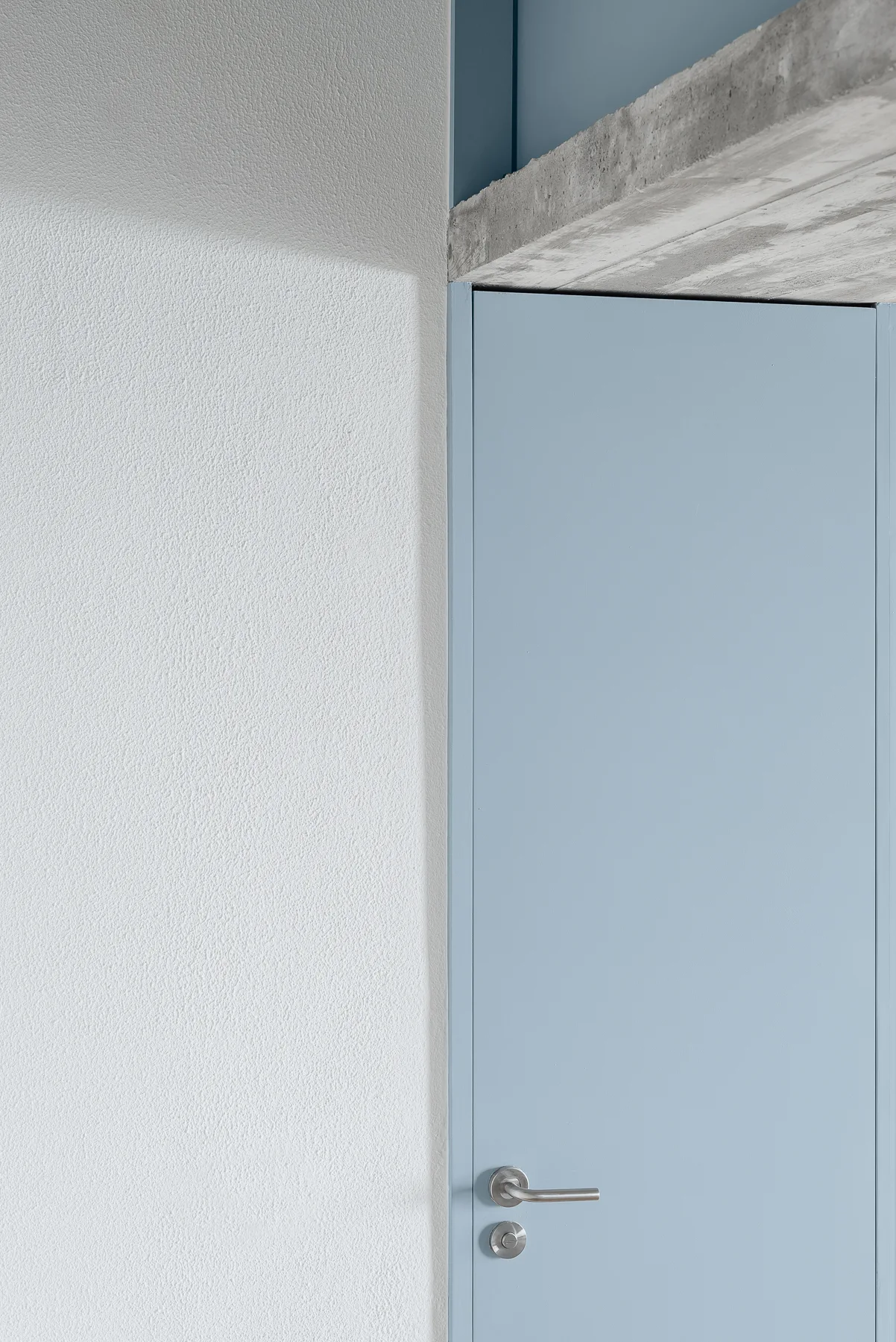 A close-up of a matte blue door with a silver handle set against a textured white plaster wall beneath an exposed concrete beam.