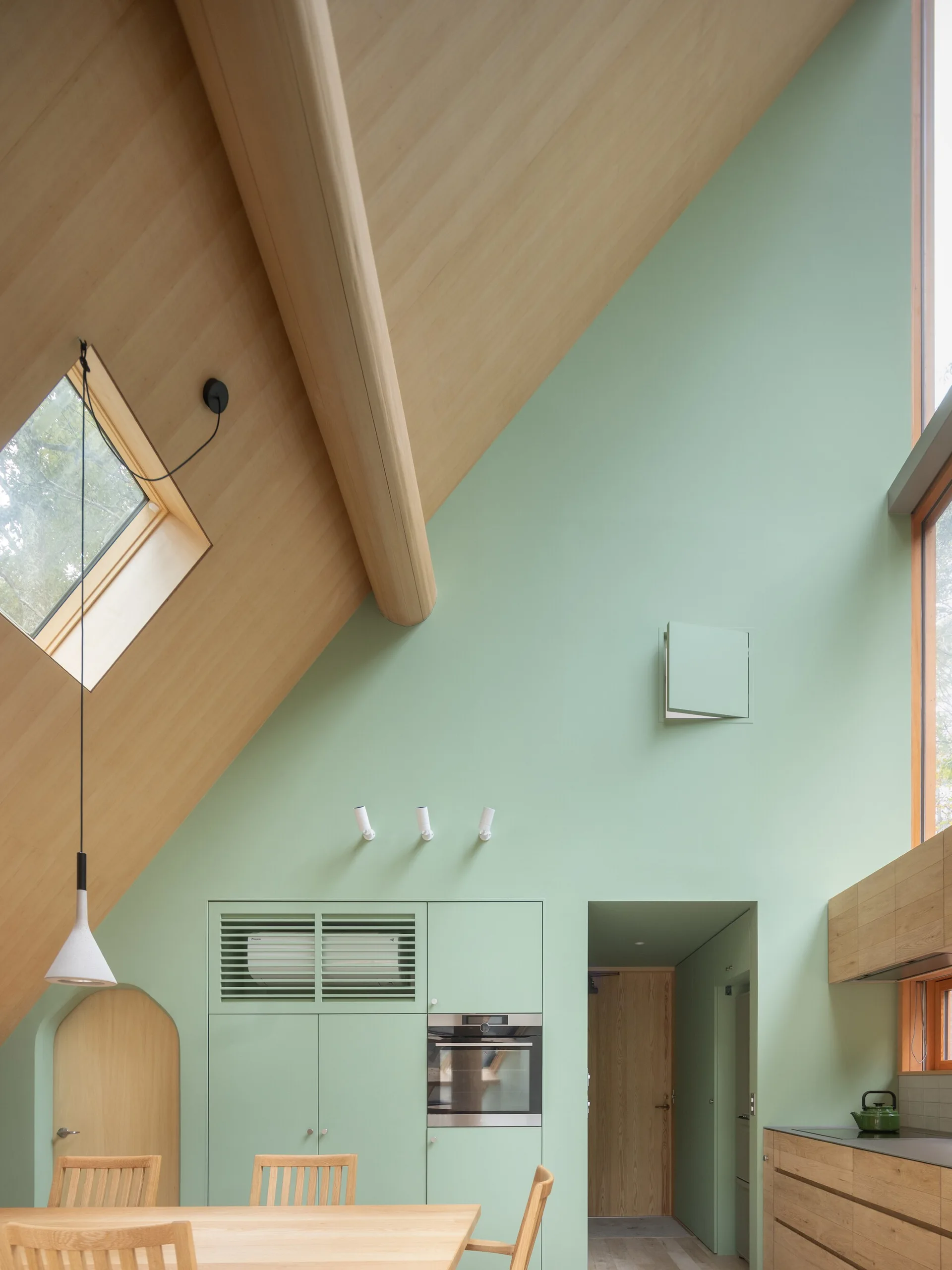 A modern kitchen features sage green walls and cabinets beneath a vaulted light wood ceiling with a skylight and wood dining table.