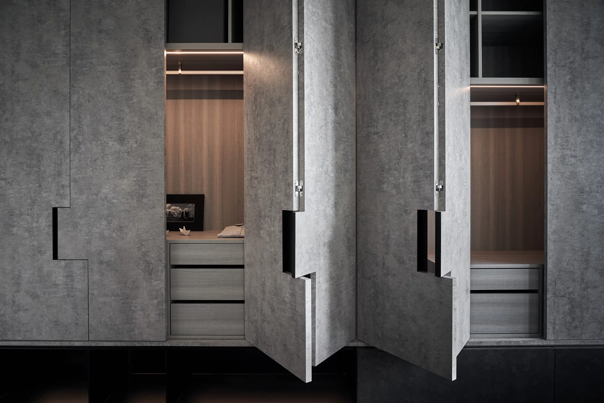 A modern wardrobe with textured grey concrete-like doors open to reveal warm wood-toned drawers, shelves, and integrated linear LED lighting.
