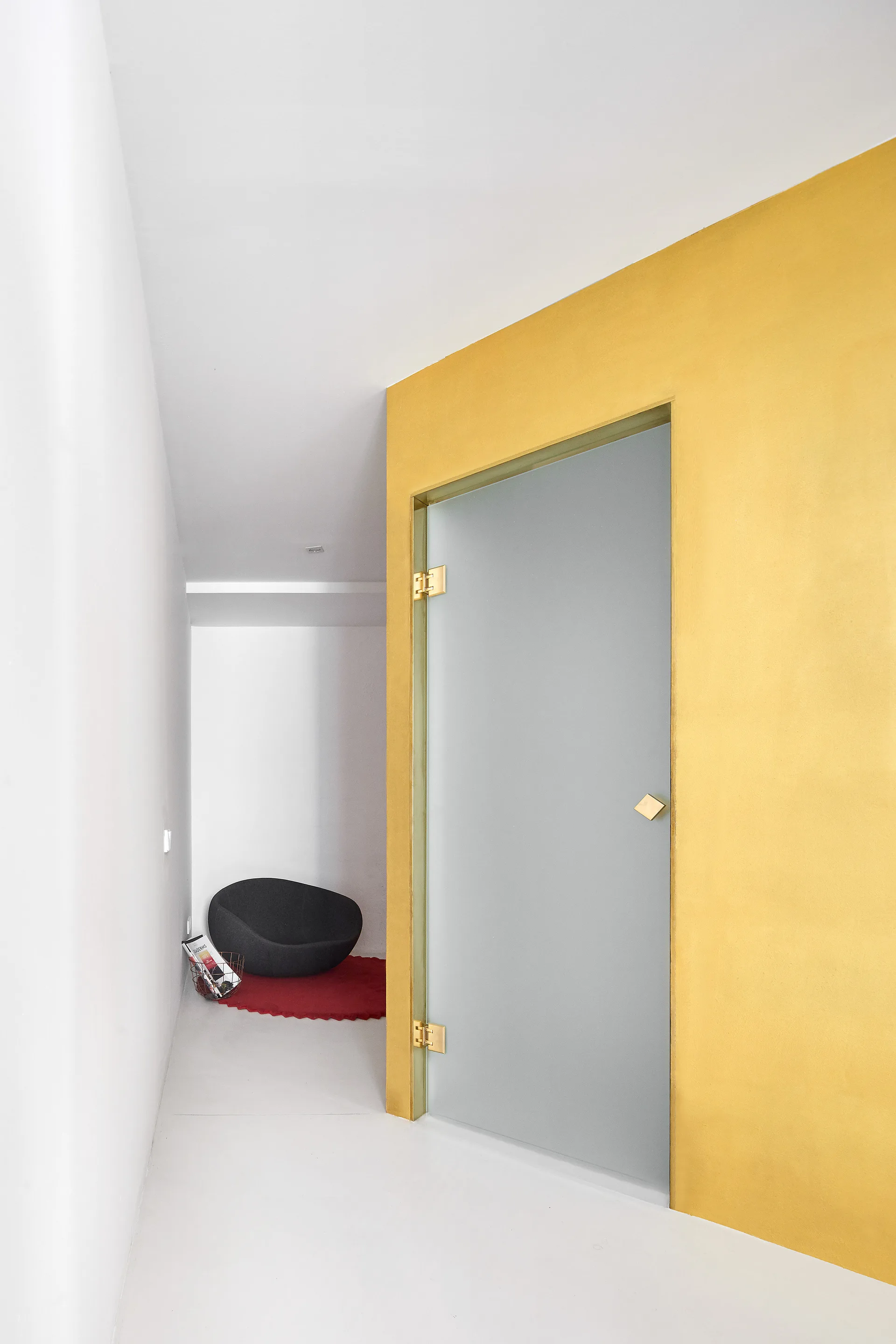 A minimalist white hallway features a bright yellow accent wall enclosing a frosted glass door with gold hinges, leading to a small reading nook.