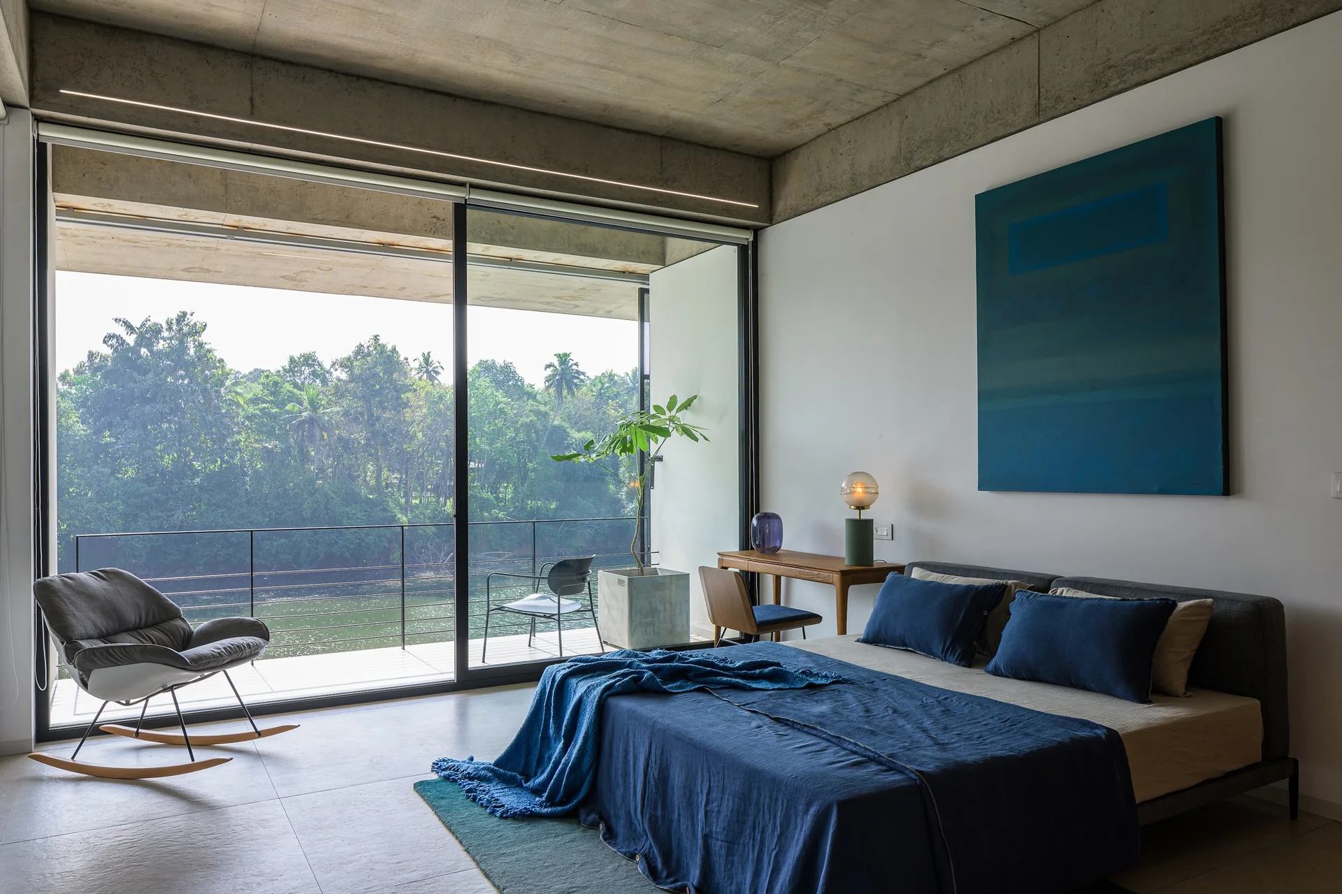 A modern bedroom features a low bed with blue linens, an exposed concrete ceiling, and sliding glass doors opening to a lush river view.
