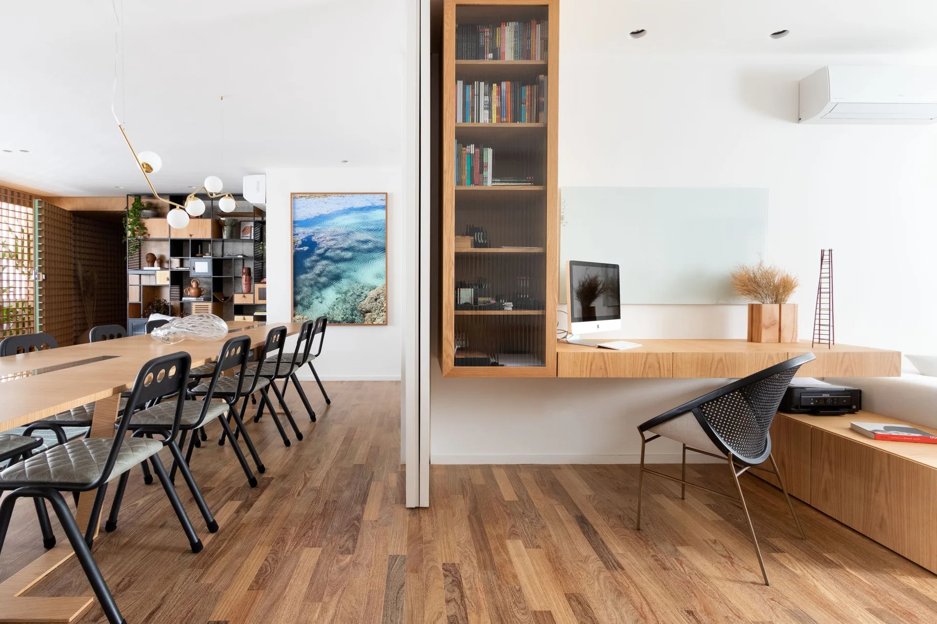 A modern open interior features a wooden dining table with black chairs separated by a partition wall from a floating desk with a tall glass bookshelf.